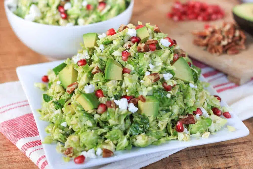 Shredded Brussels Sprout Pomegranate Salad with Honey Mustard Dressing - Served on a Plate with Another Bowl Full of the Delicious Dish in the Background