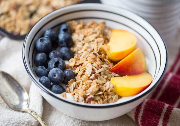 Coconut Maple Pecan Granola Beauty Shot with a Full Bowl of Granola, Berries and Peaches, and a Spoon Right Next to It