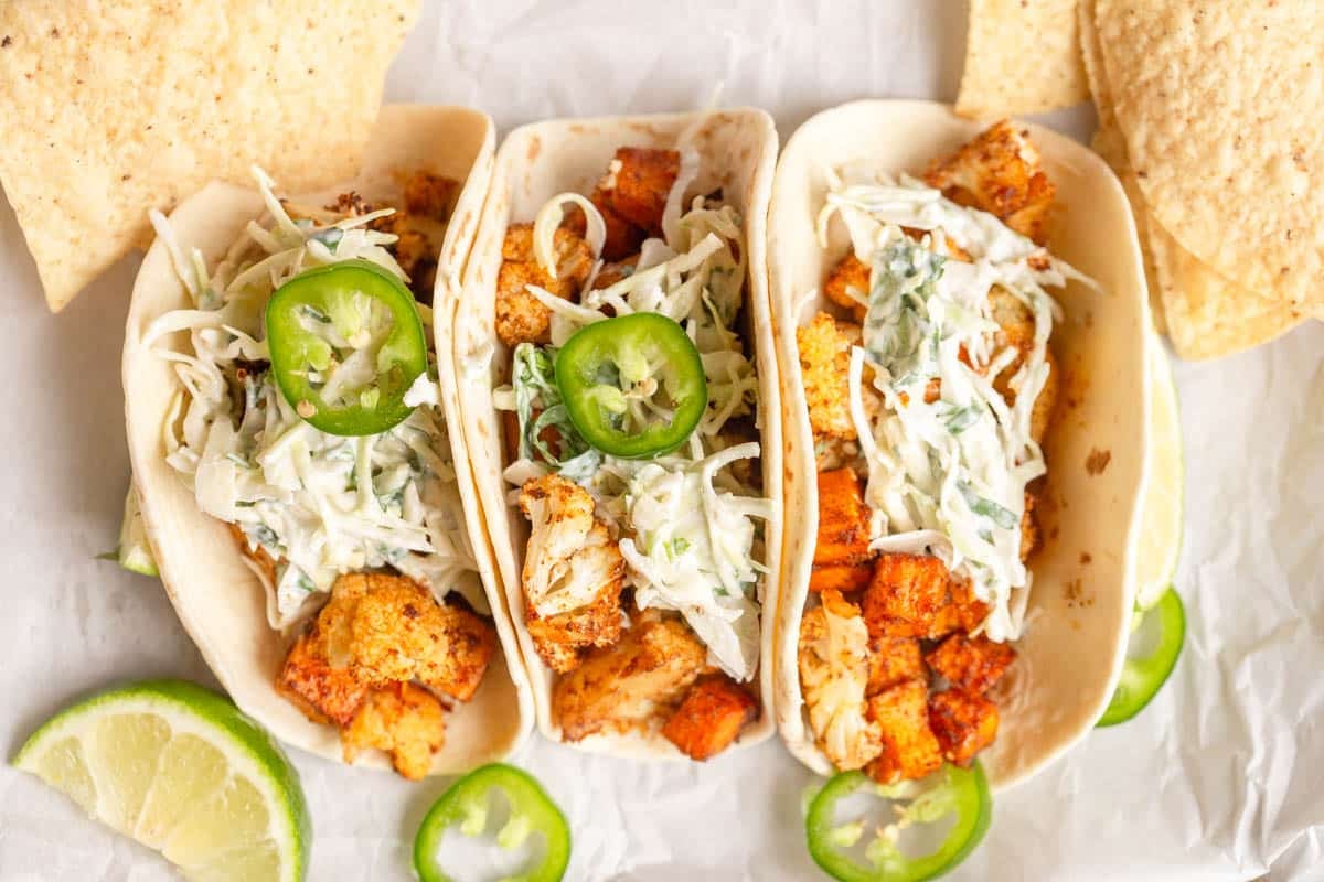3 Roasted Sweet Potato Cauliflower Tacos on a piece of parchment paper with lime wedges, jalapeno slices, and tortilla chips around them.