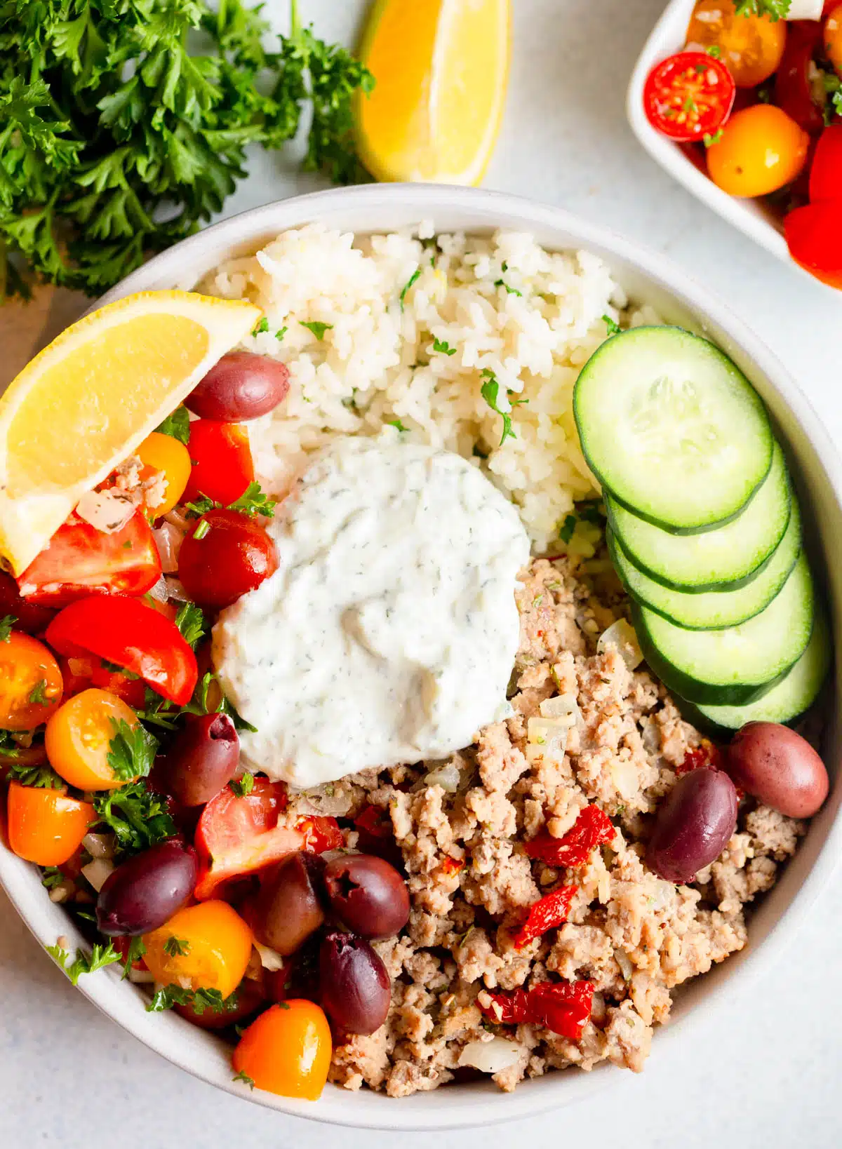 Greek Turkey Rice Bowls in a shallow white bowl with tzatziki on top, tomato salad and fresh parsley in the background. 