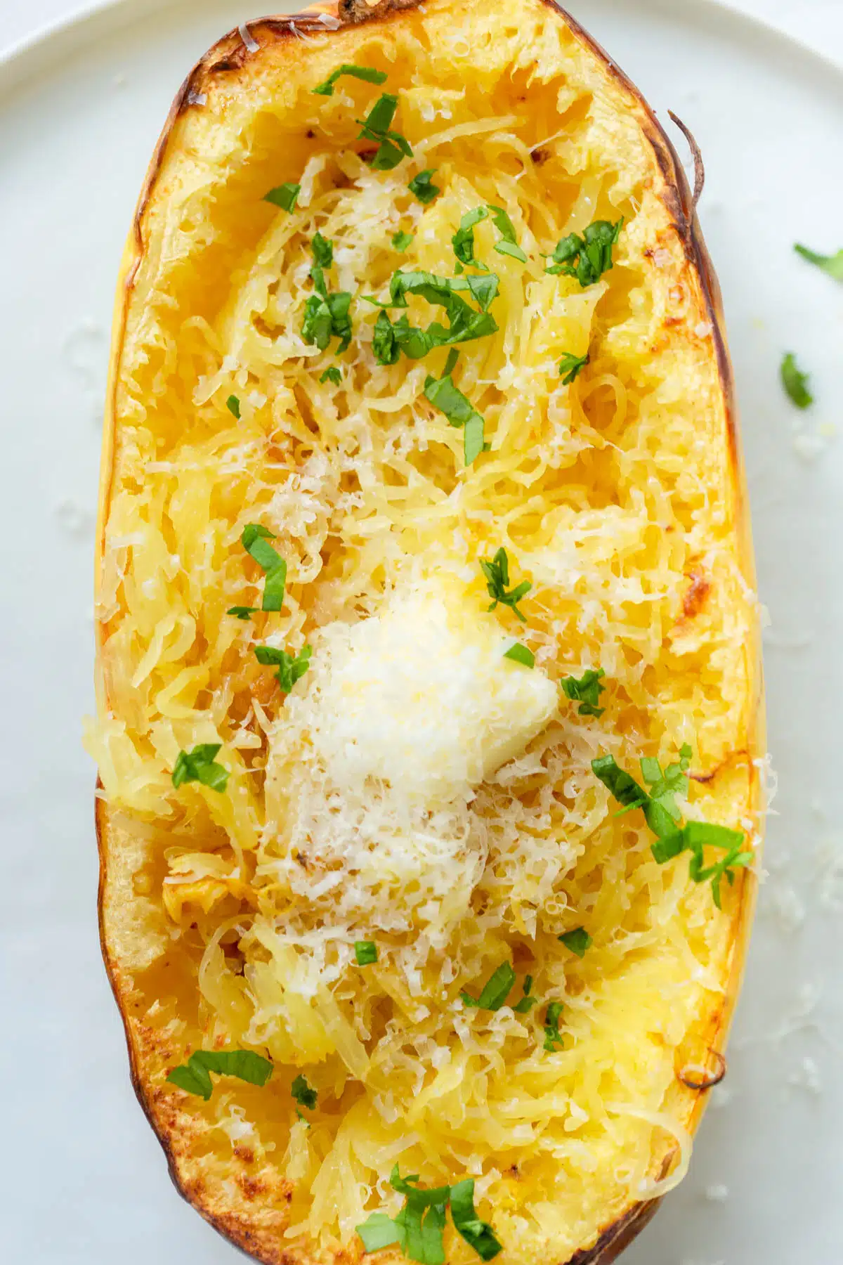 One half of a spaghetti squash on a white plate butter, Parmesan cheese, and chopped herbs on top. 