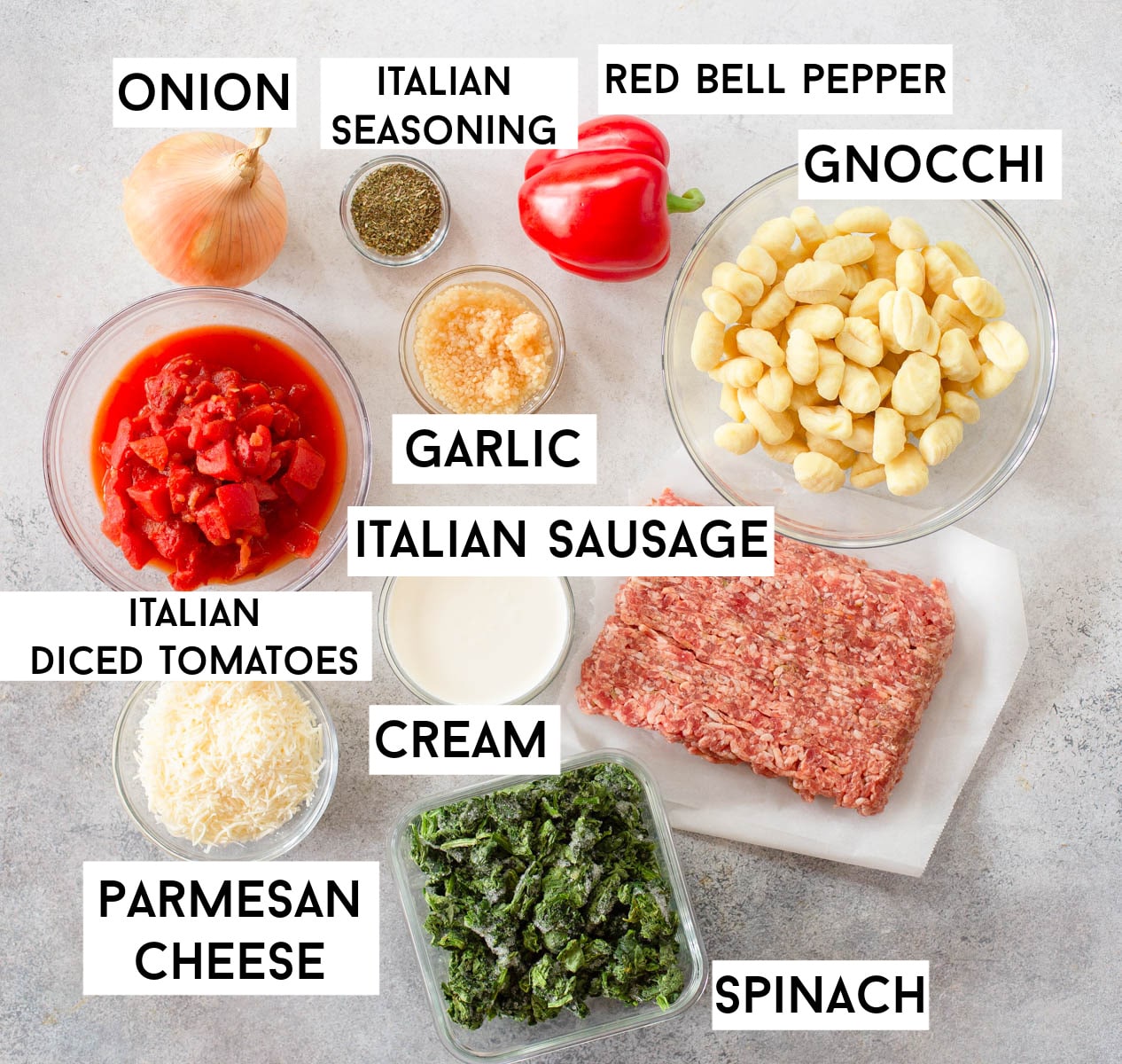Ingredients in Italian Sausage Gnocchi Soup laid out on a gray background including onion, Italian seasoning, red bell pepper, fresh garlic, gnocchi, Italian diced tomatoes, Italian sausage, cream, Parmesan cheese, and spinach.