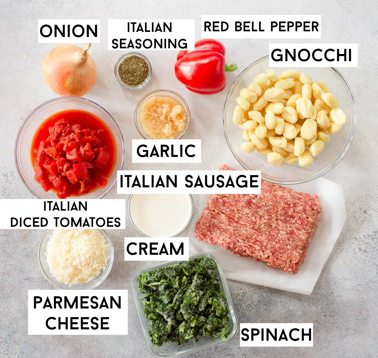 Ingredients in Italian Sausage Gnocchi Soup laid out on a gray background including onion, Italian seasoning, red bell pepper, fresh garlic, gnocchi, Italian diced tomatoes, Italian sausage, cream, Parmesan cheese, and spinach. 