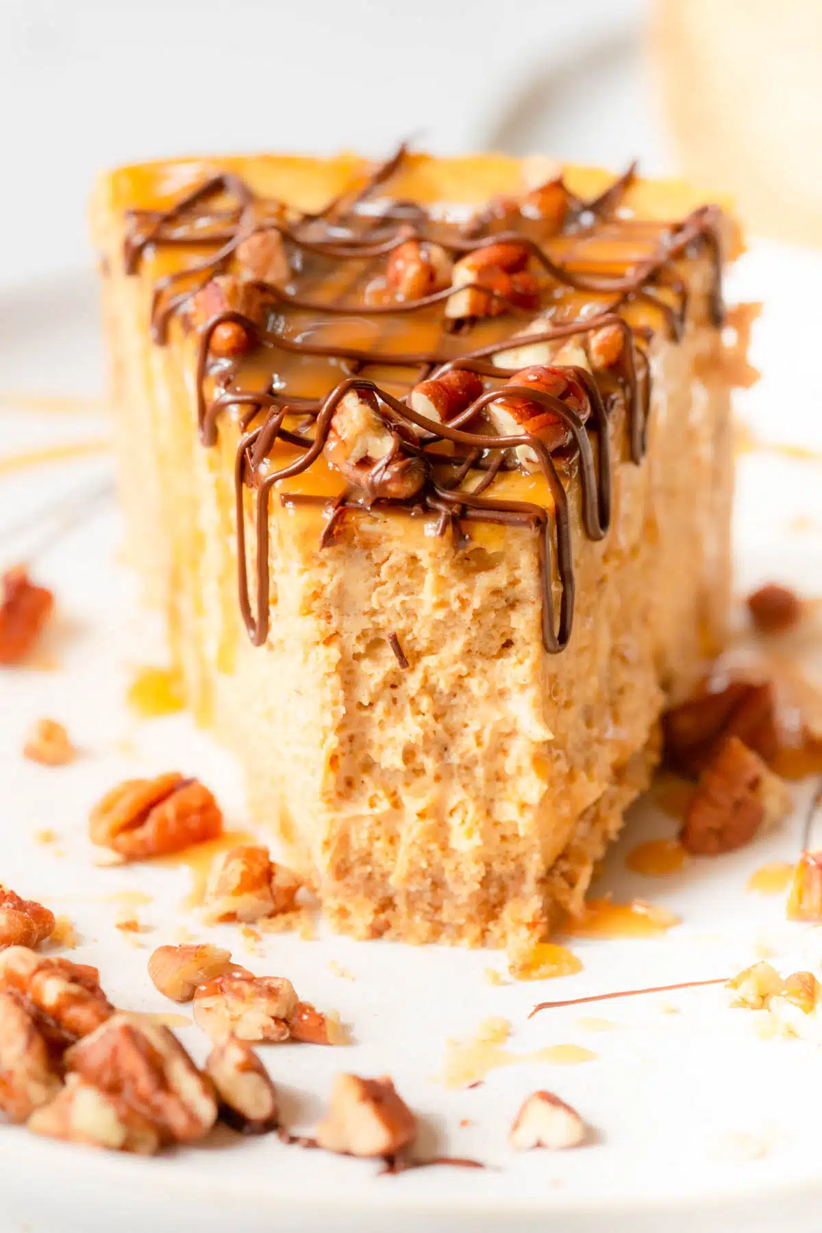 A slice of Turtle Pumpkin Cheesecake on a white plate with chopped pecans scattered around the plate. A forkful has been taken out of the front of the slice of cheesecake. 