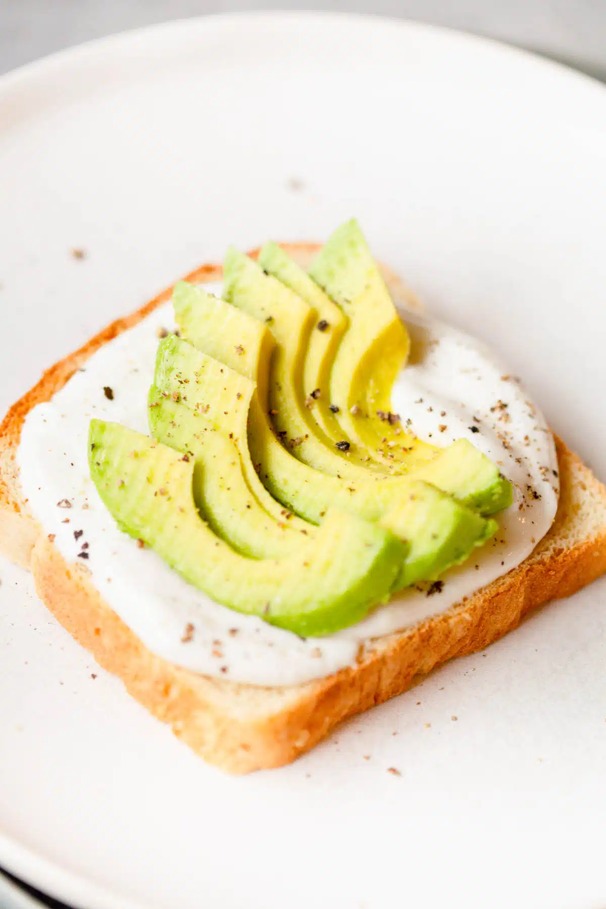 Cottage cheese toast with sliced avocado and fresh cracked pepper on top, on a white plate. 