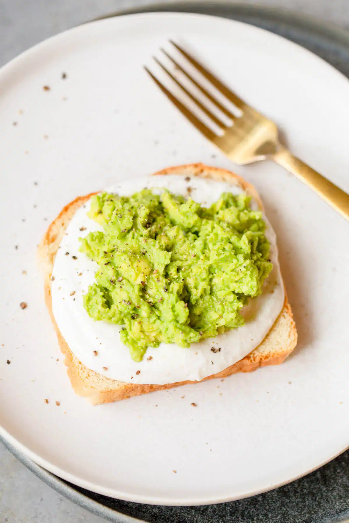 Avocado Cottage Cheese Toast on an ivory plate topped with fresh cracked pepper and a gold fork in the background. 
