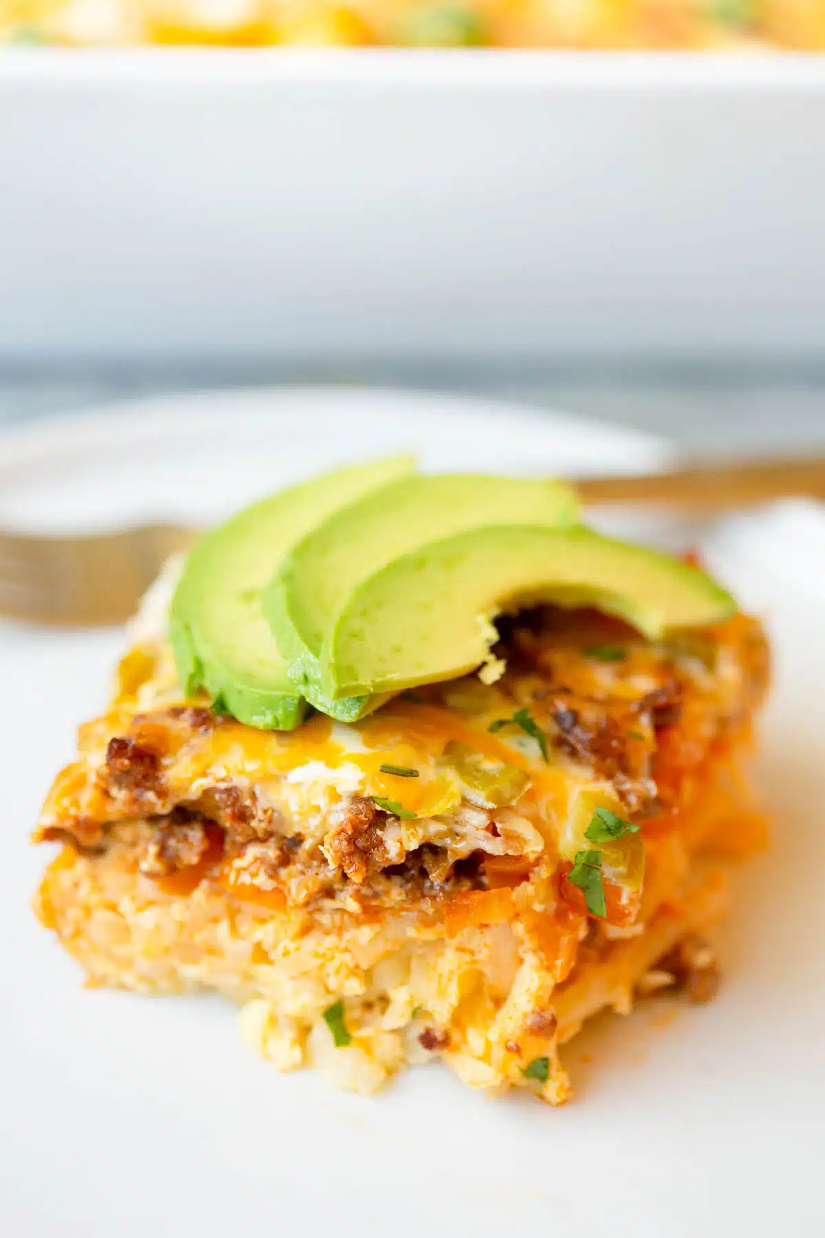 A square slice of Chorizo Breakfast Casserole on a white plate with avocado slices and chopped cilantro on top.