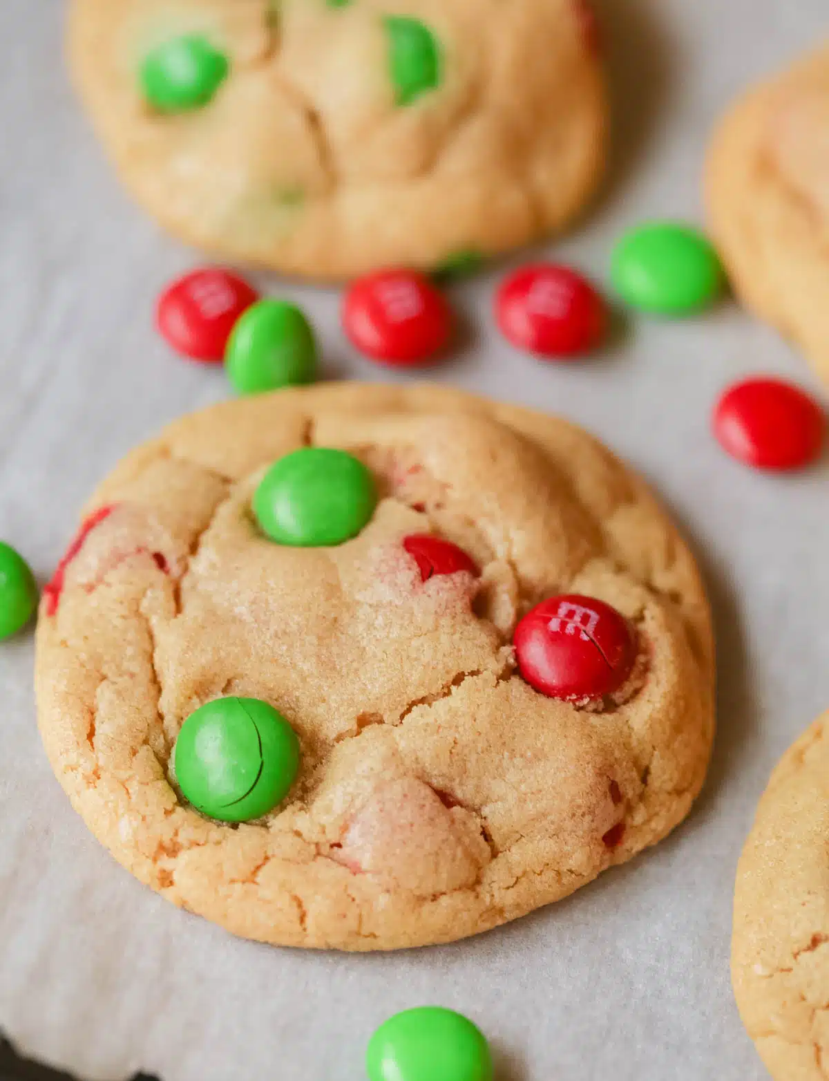 A Christmas Peanut Butter M&M Cookie on parchment paper with Christmas colored M&Ms scattered around it. 