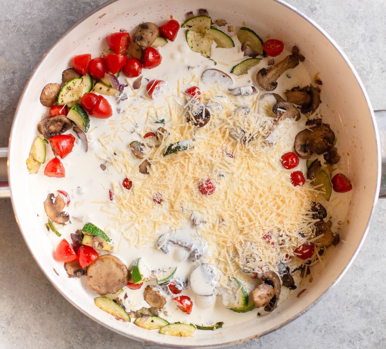 White pan with vegetables in it along with heavy cream and shredded Parmesan on top.
