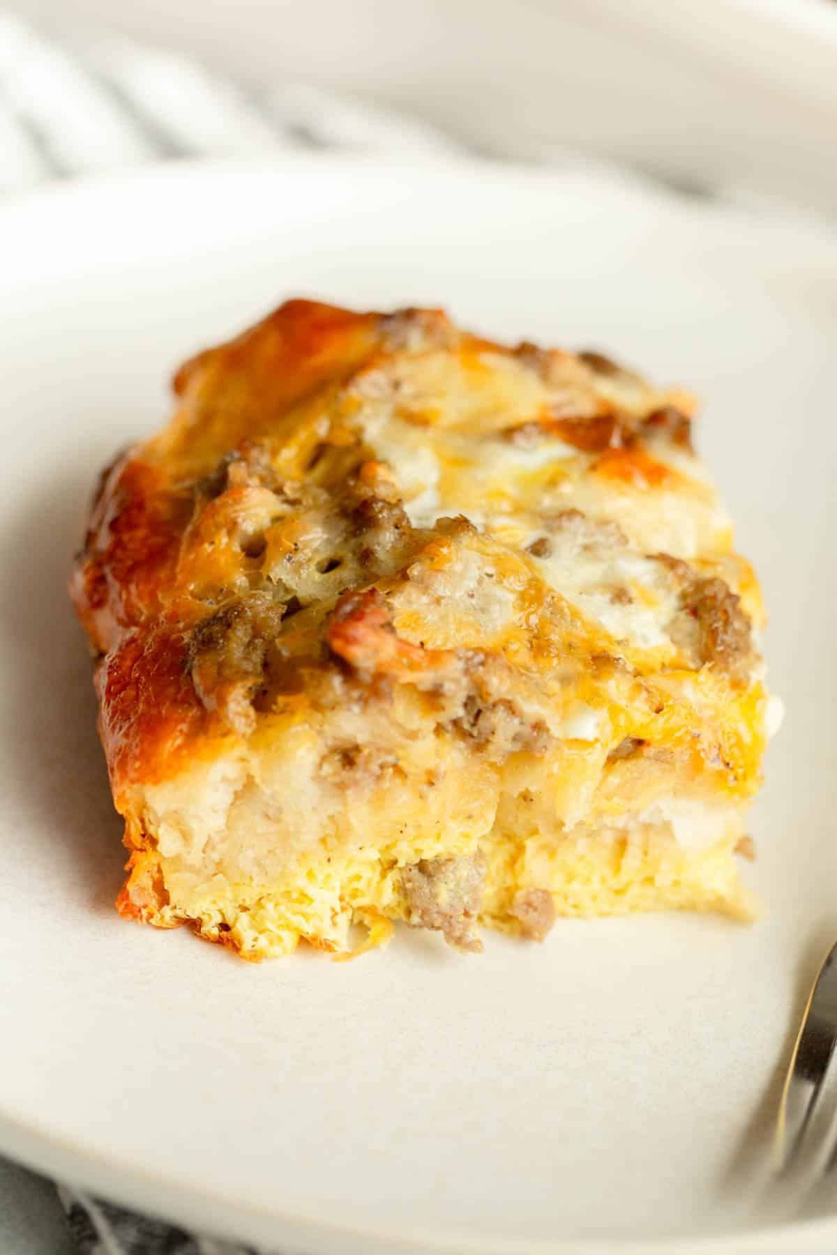 A piece of Sausage Egg Cheese Biscuit Casserole on a cream colored plate with a silver fork next to it.