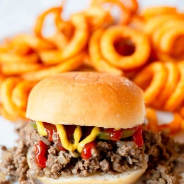 Maid Rite Sandwich topped with pickles, ketchup, and mustard, with fries in the background.