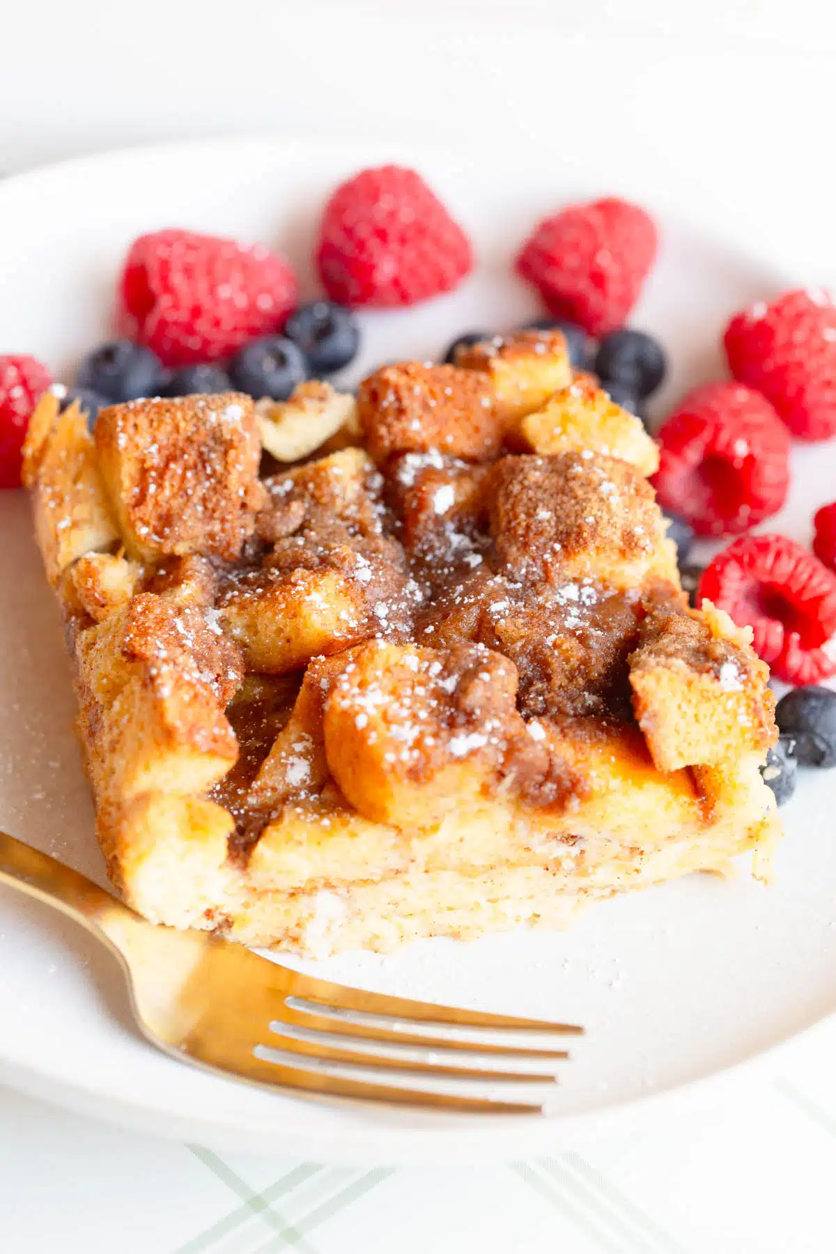 A slice of Brioche French Toast Casserole topped with powdered sugar and maple syrup, on a white plate with raspberries and blueberries in the background and a gold fork next to it. 