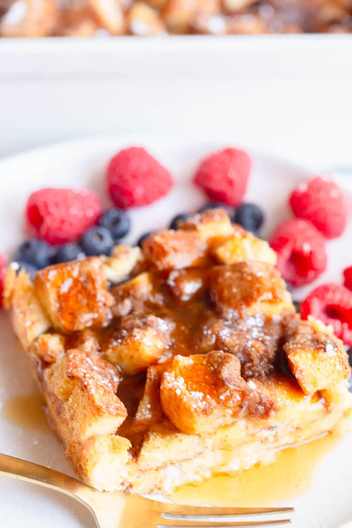 A slice of Brioche French Toast Casserole topped with powdered sugar and maple syrup, on a white plate with raspberries and blueberries in the background and a gold fork next to it. 