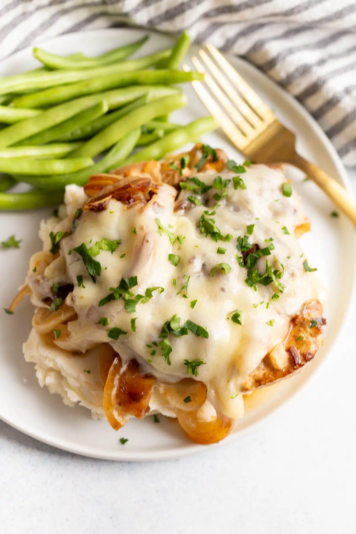 Slow Cooker French Onion Chicken on top of mashed potatoes on a white plate with a gold fork and green beans in the background.