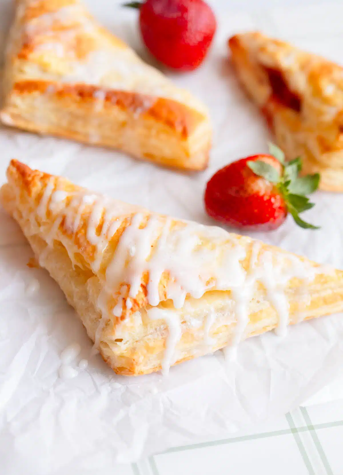 Strawberry Turnovers drizzled with a powdered sugar glaze on parchment paper with fresh strawberries in the background.