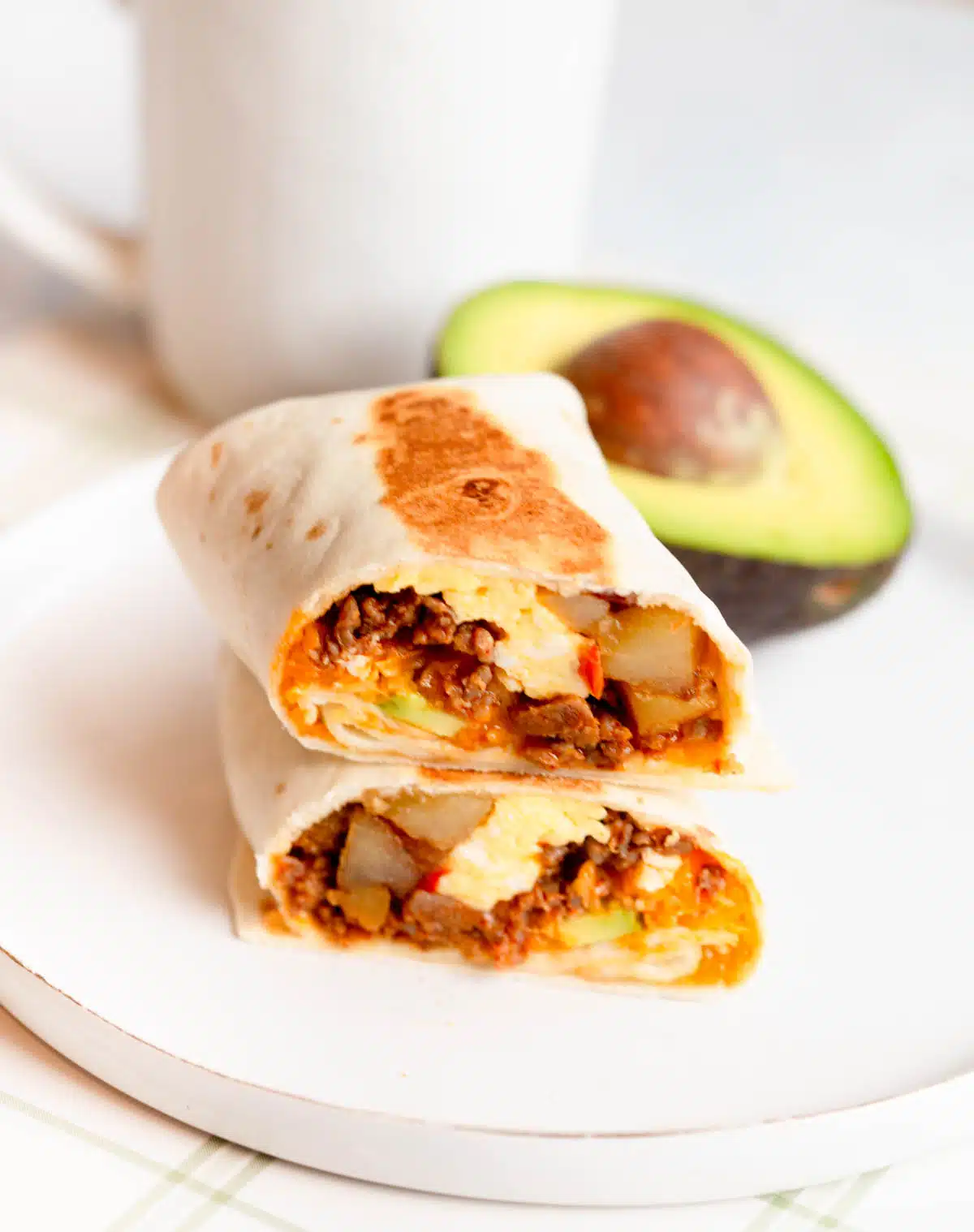 Chorizo Potato Breakfast Burrito cut in half with one half sitting on top of the other one, with an avocado in the background.