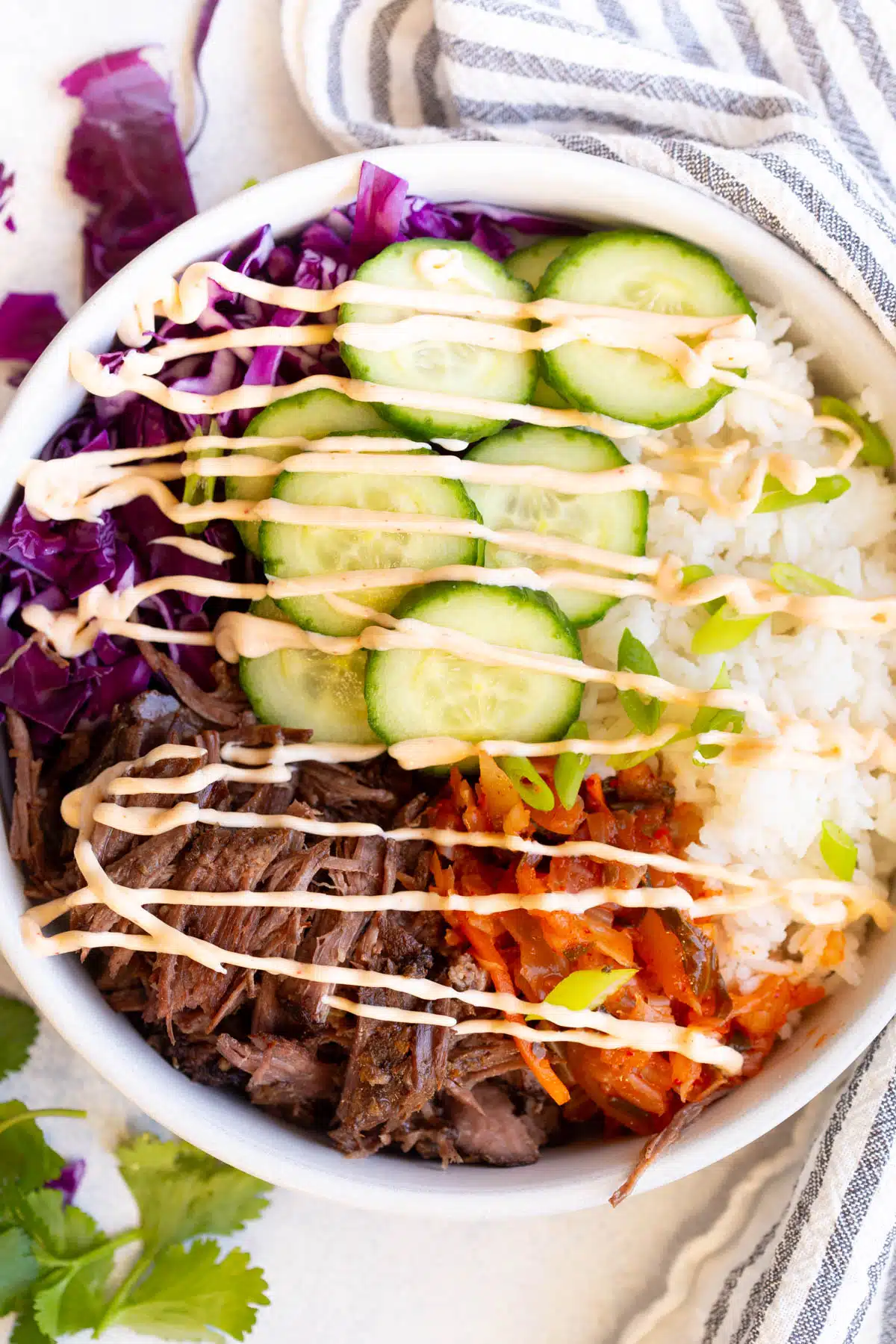 Slow Cooker Korean Beef Burrito Bowls in a shallow white bowl with a blue and white striped towel wrapped around it and some cabbage and cilantro on the gray background.