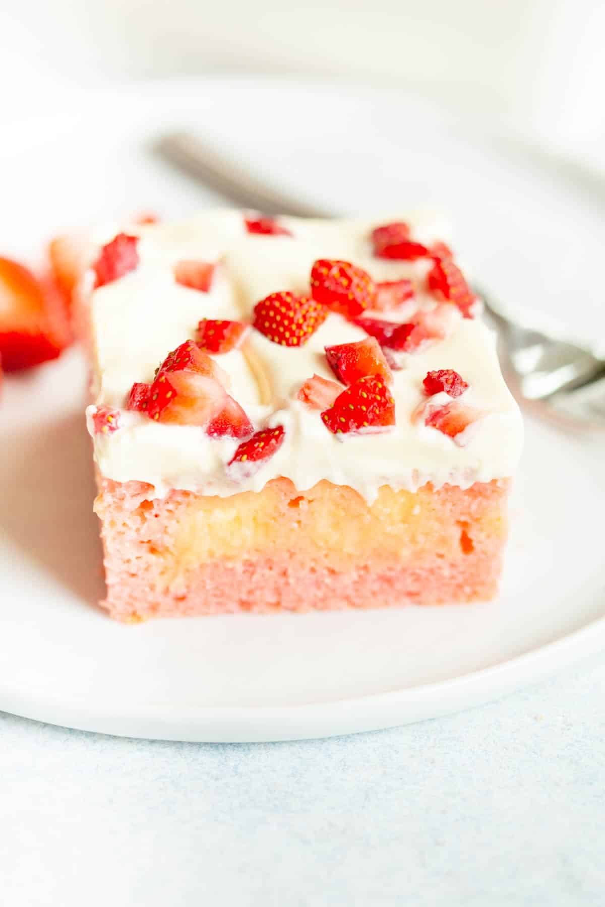 A slice of Strawberry Pudding Poke Cake on a white plate, topped with chopped strawberries. There is a silver fork in the background.