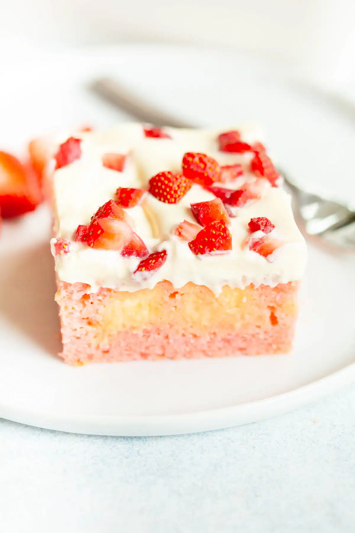 A slice of Strawberry Pudding Poke Cake on a white plate, topped with chopped strawberries. There is a silver fork in the background.