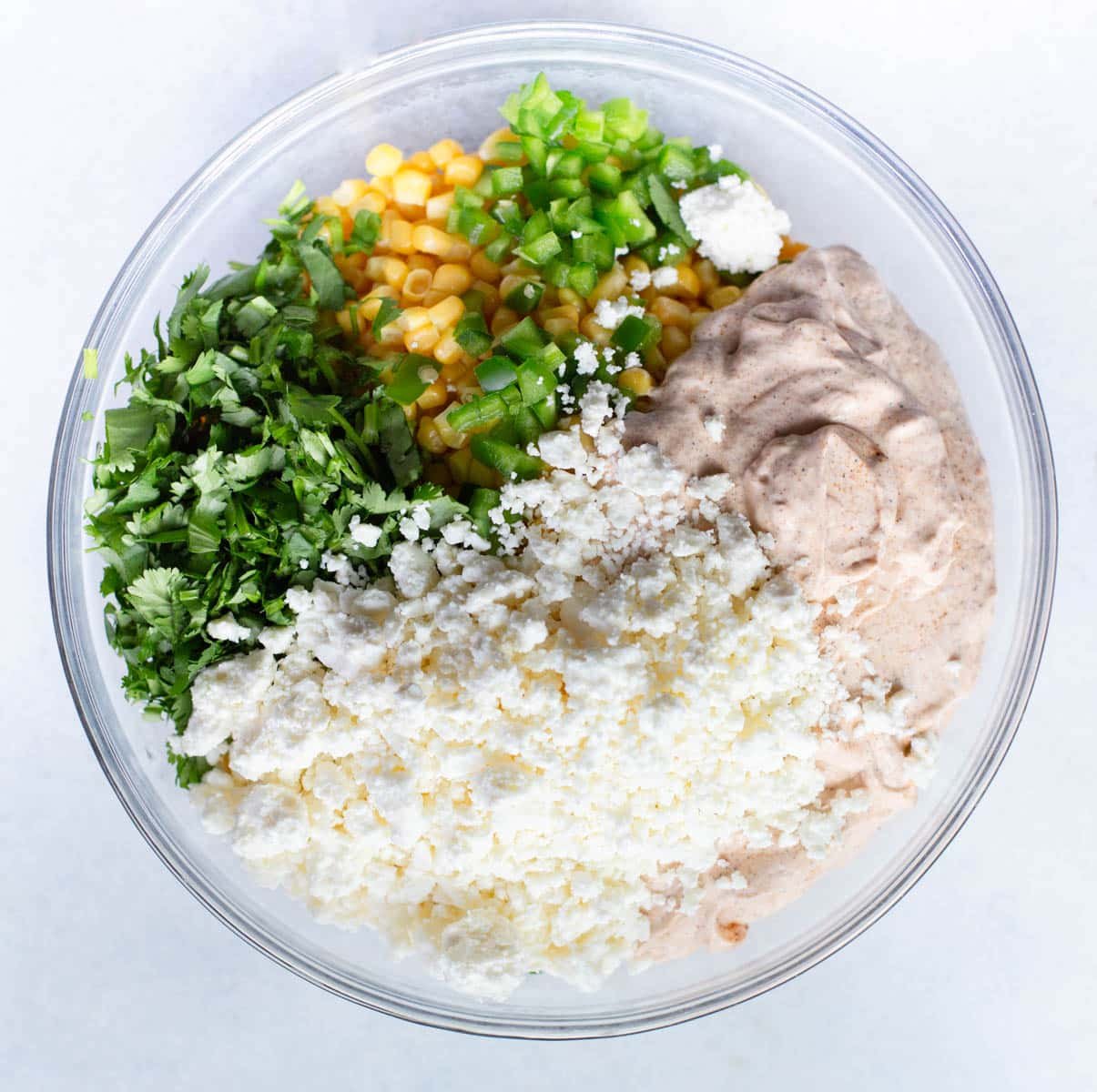 Glass bowl with corn, jalapeño, cilantro, feta cheese, and street corn dip sauce in it with a gray background.