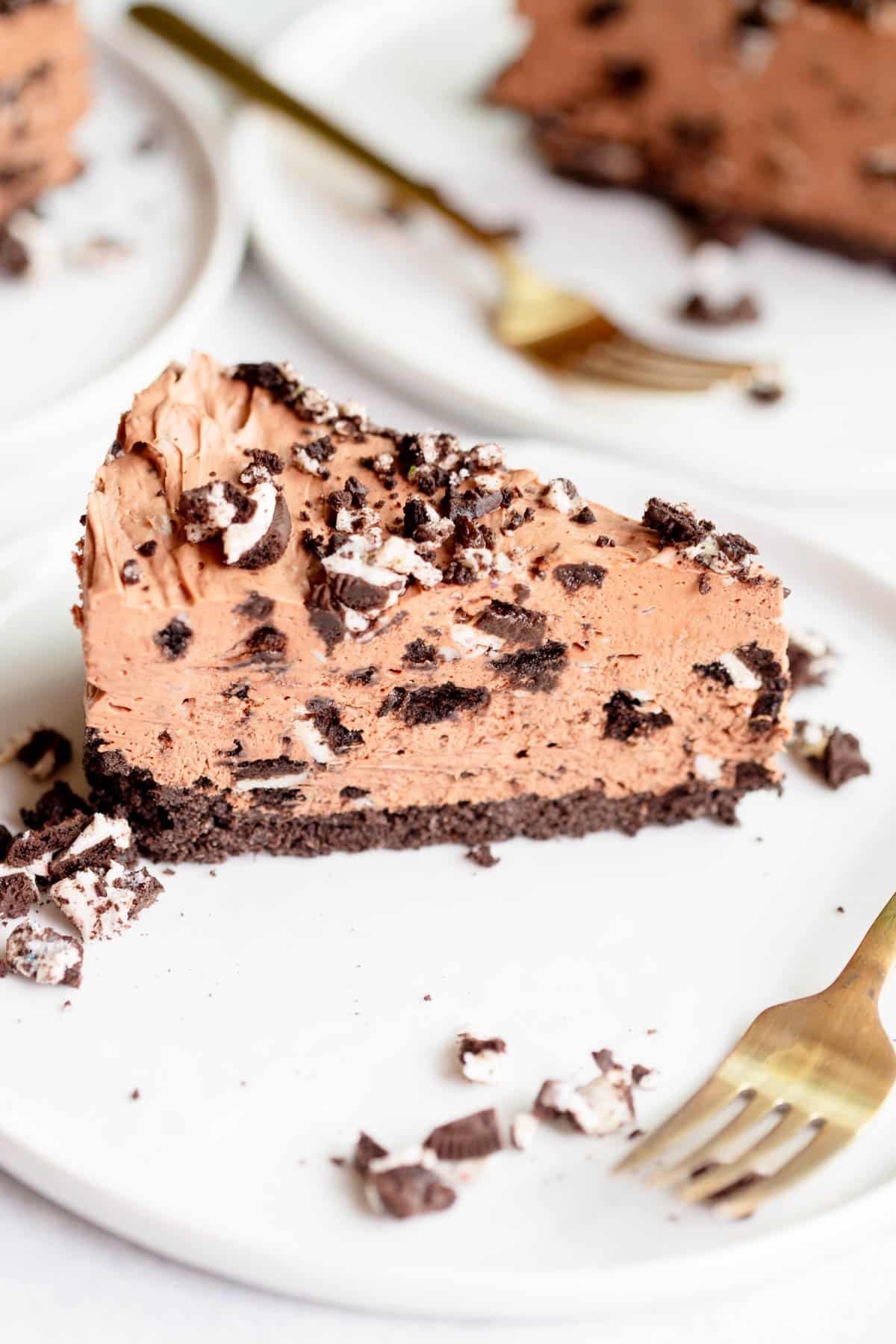 A slice of no Bake Oreo Cheesecake on a white plate, topped with chopped Oreos and more chopped Oreos scattered on the plate. In the background there is another slice of No Bake Oreo Cheesecake with a gold fork next to it.