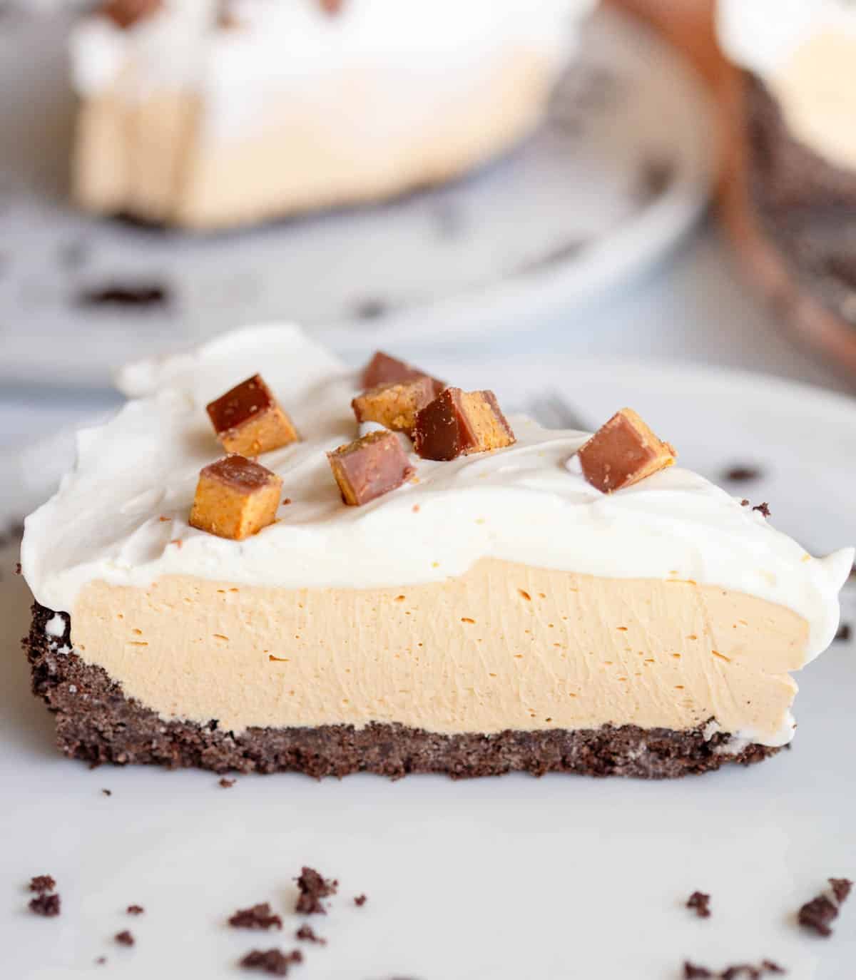 A slice of No Bake Peanut Butter Pie topped with diced peanut butter cups on a white plate with Oreo crumb scattered around it and another slice blurred in the background.