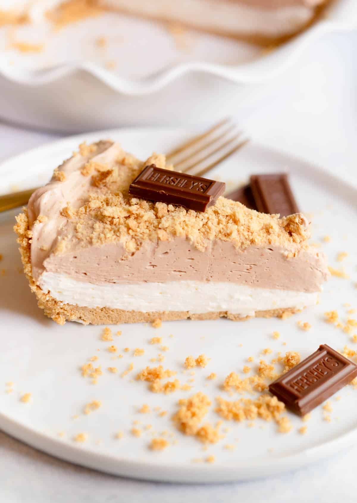 A slice of No Bake S'Mores Pie on a white plate topped with graham cracker crumbs and a slice of Hershey's chocolate. 