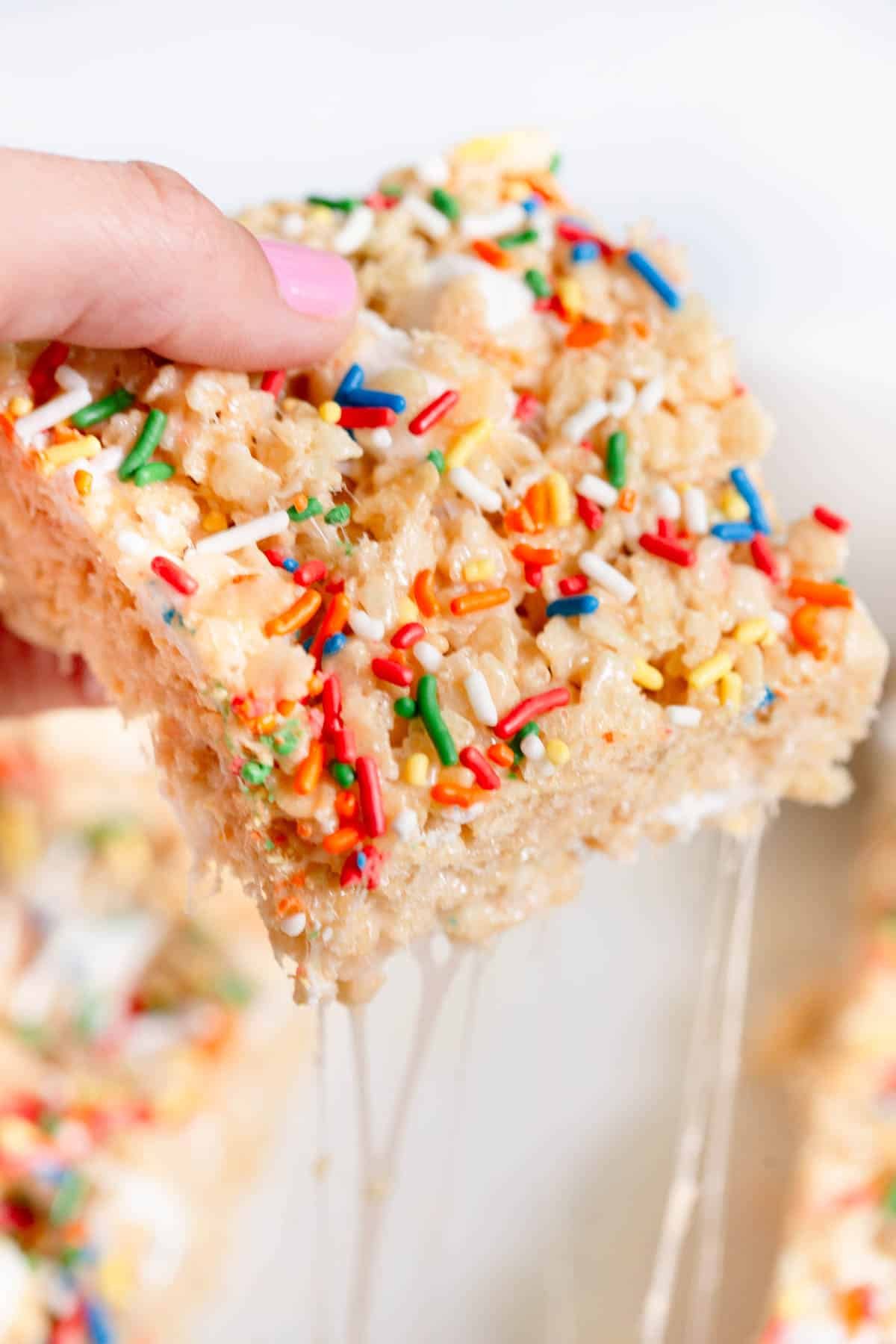 A hand with pink fingernails hold a Birthday Cake Rice Krispie Treat that has gooey marshmallow sticking to the other piece. 