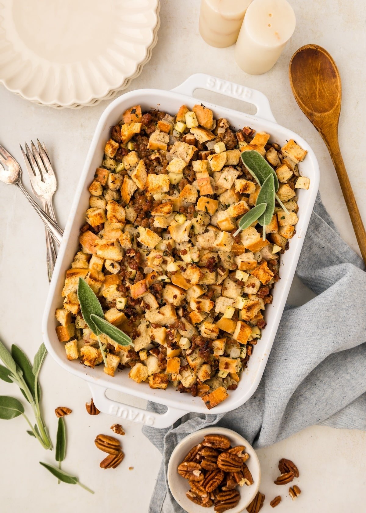 A white Staub baking dish with Sausage Pecan Stuffing with fresh sage leaves on top as garnish. There is more fresh sage, three vintage forks, pecans, a wooden spoon, scalloped plates, salt and pepper shakers, and a gray towel around the baking dish. 