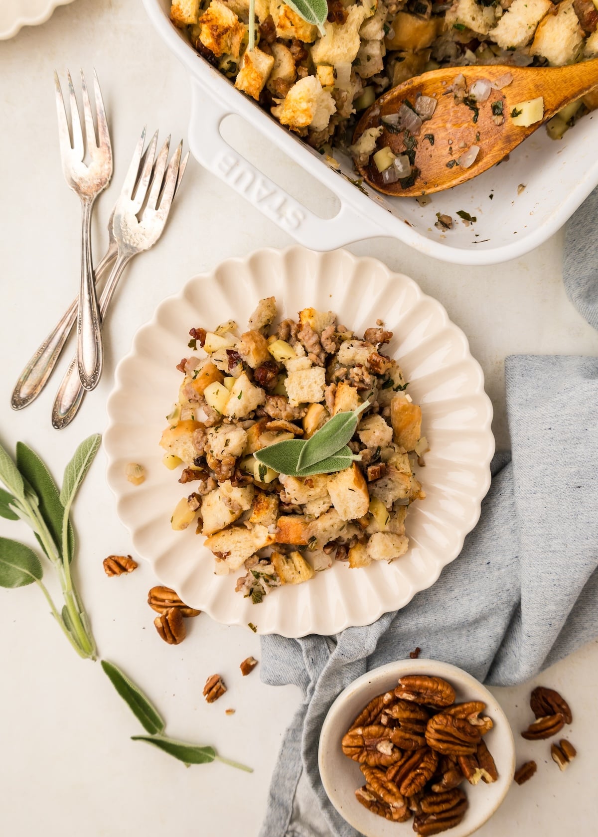 Sausage Pecan Stuffing on a scalloped cream color plate with two fresh sage leaves on top as garnish. There are three vintage forks next to the plate along with more fish sage, pecans, and a gray towel. 