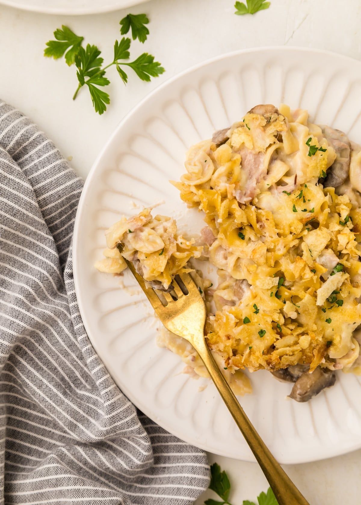 Tuna Noodle Casserole with Potato Chips on a white scalloped plate, with a gold fork, and a gray striped napkin next to it. 