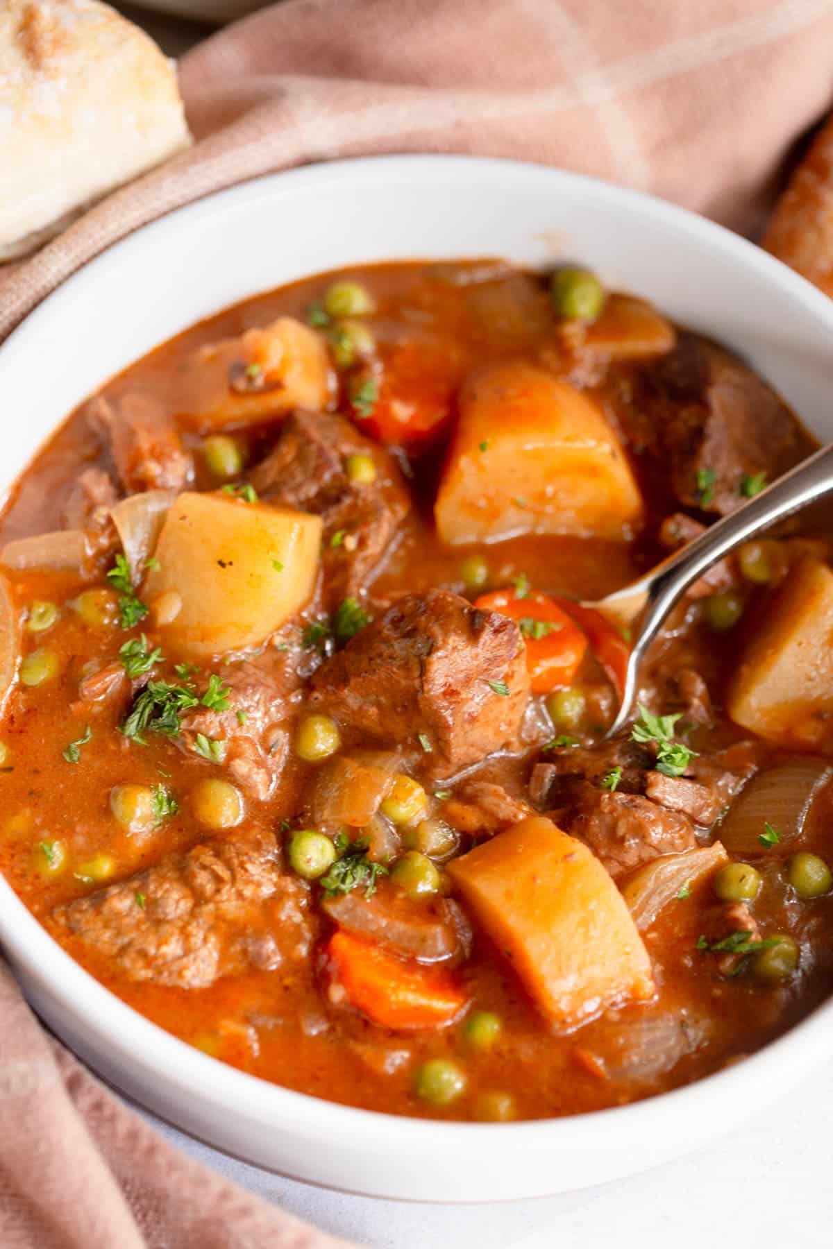 Slow Cooker Beef Stew in a large white bowl with spoon in it, and a terra cotta colored towel behind it. 