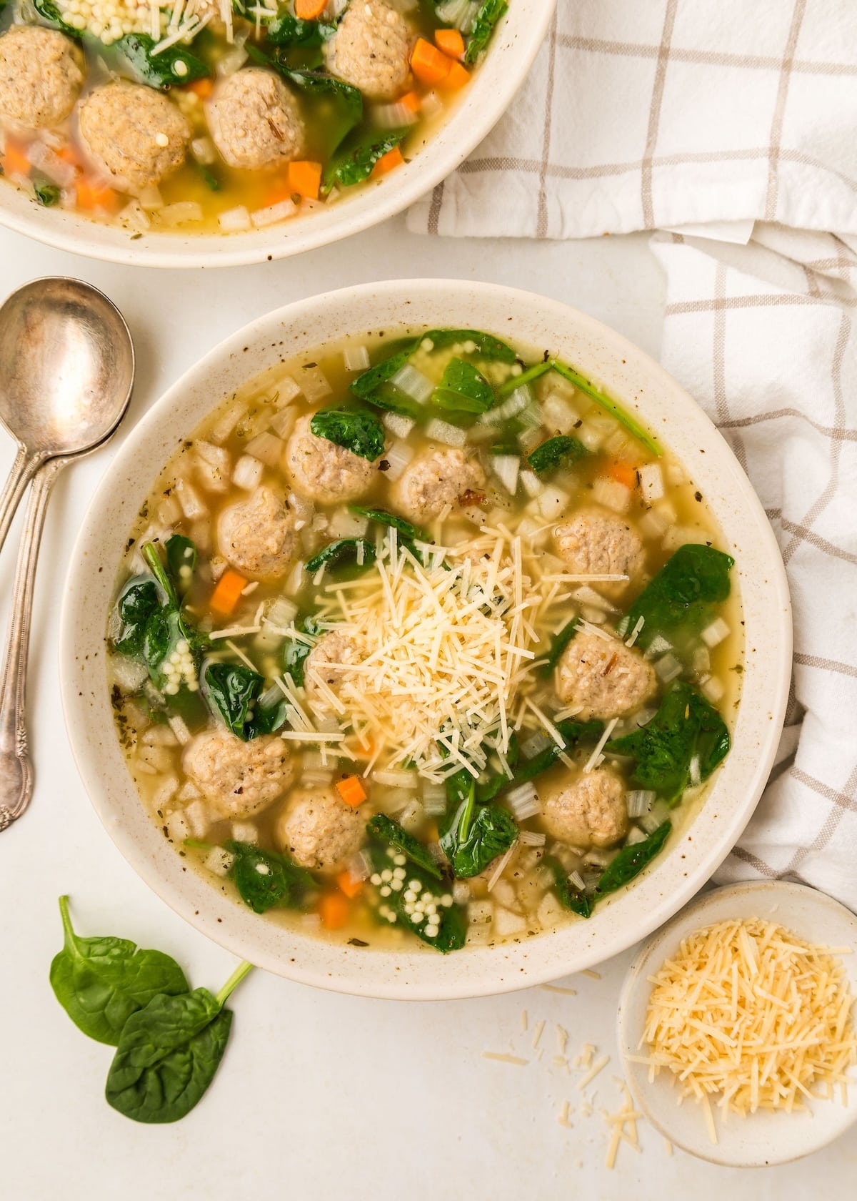 Slow Cooker Italian Wedding Soup in a speckled cream colored bowl with two antique spoons next to it along with a plaid napkin. 