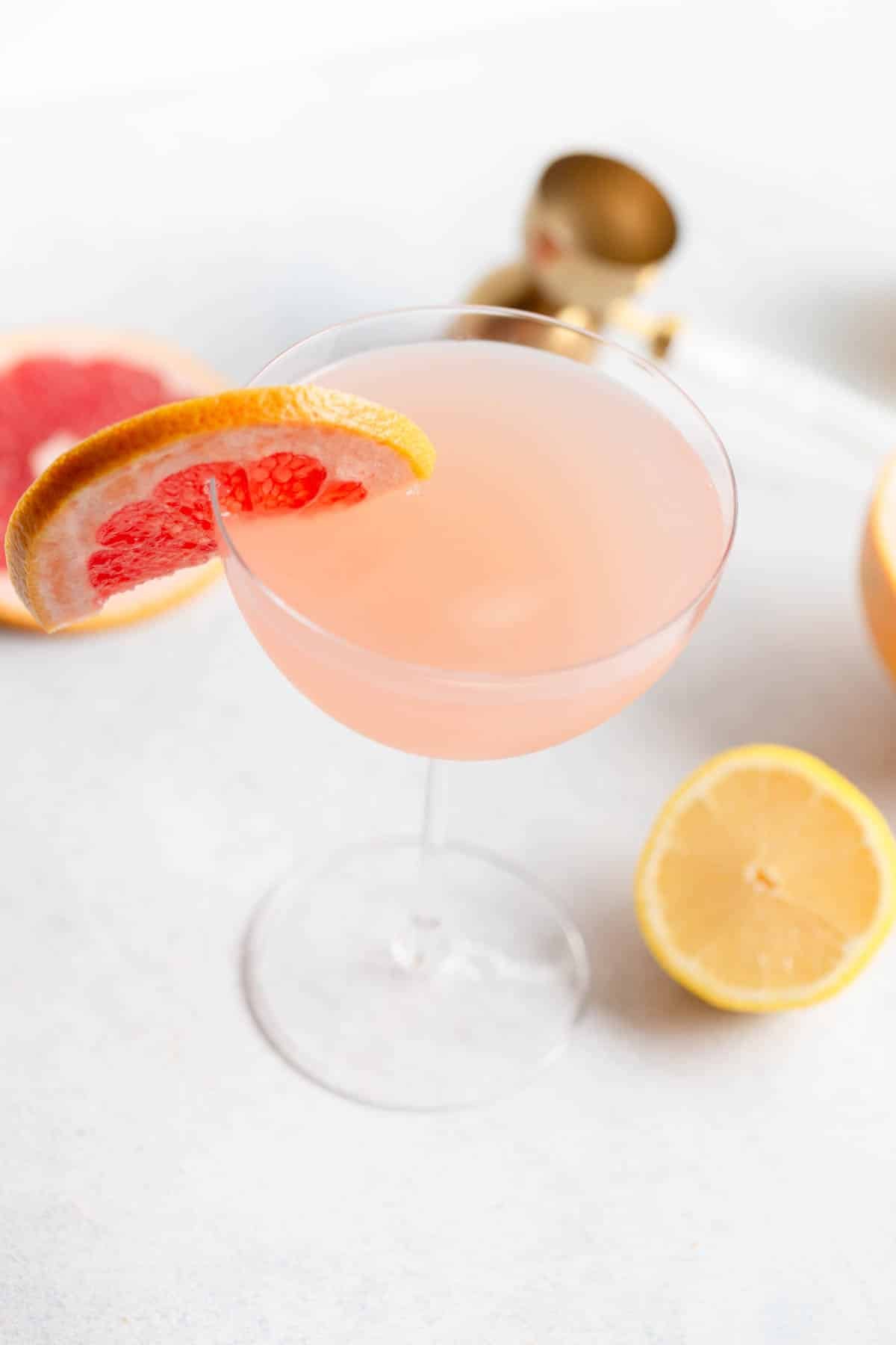 Grapefruit Martini in a martini glass with a slice of grapefruit on the rim. There is a gold shot measure tool behind it along with slices of grapefruit and lemon. 