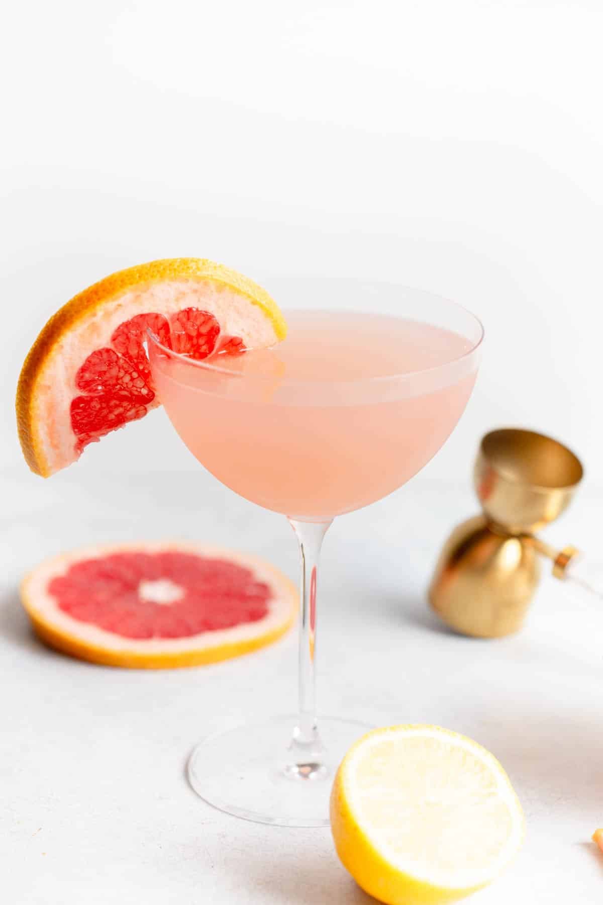 Grapefruit Martini in a martini glass with a slice of grapefruit on the rim. There is a gold shot measure tool behind it along with slices of grapefruit and lemon. 