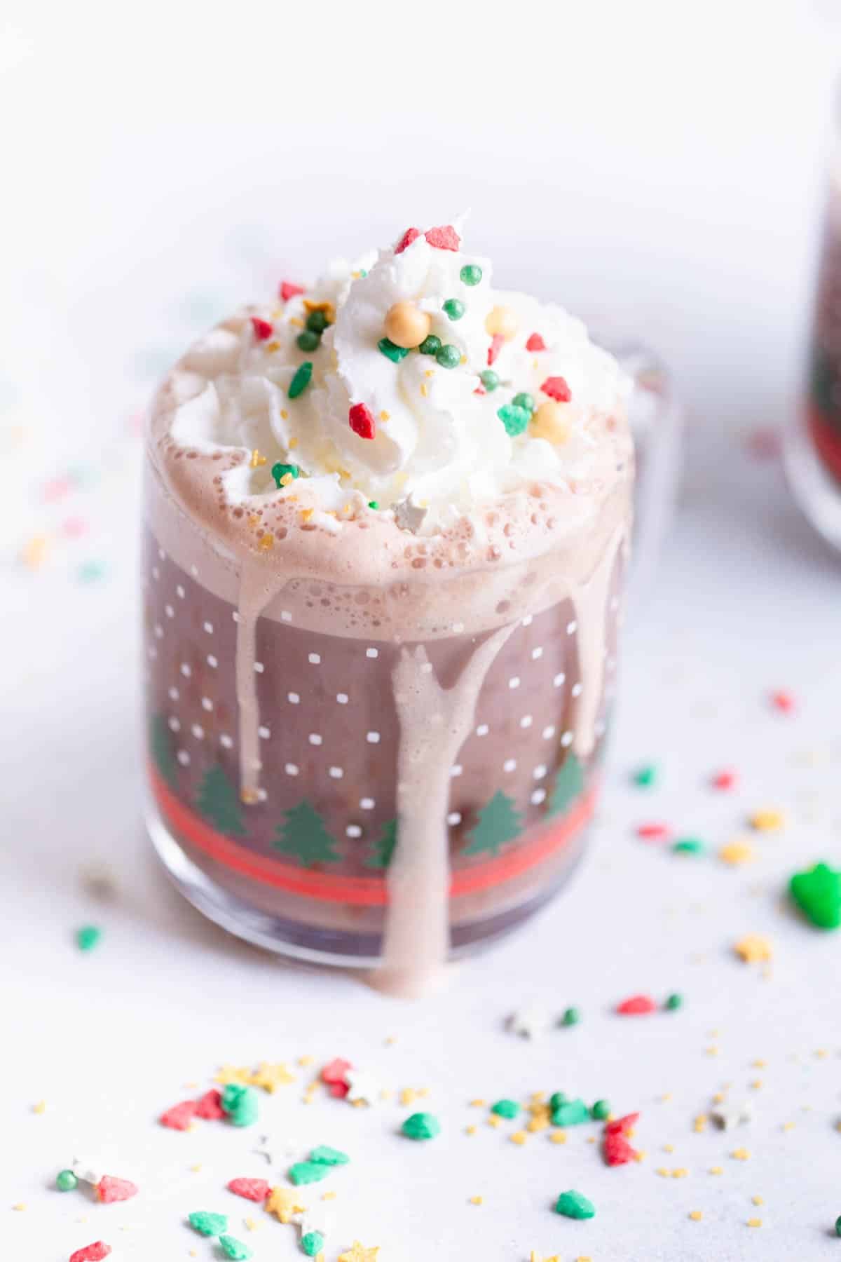 Homemade Hot Cocoa Mix, prepared, in a festive clear mug with Christmas trees on it. The hot cocoa has whipped cream on top with Christmas sprinkles on top and around the mug. 