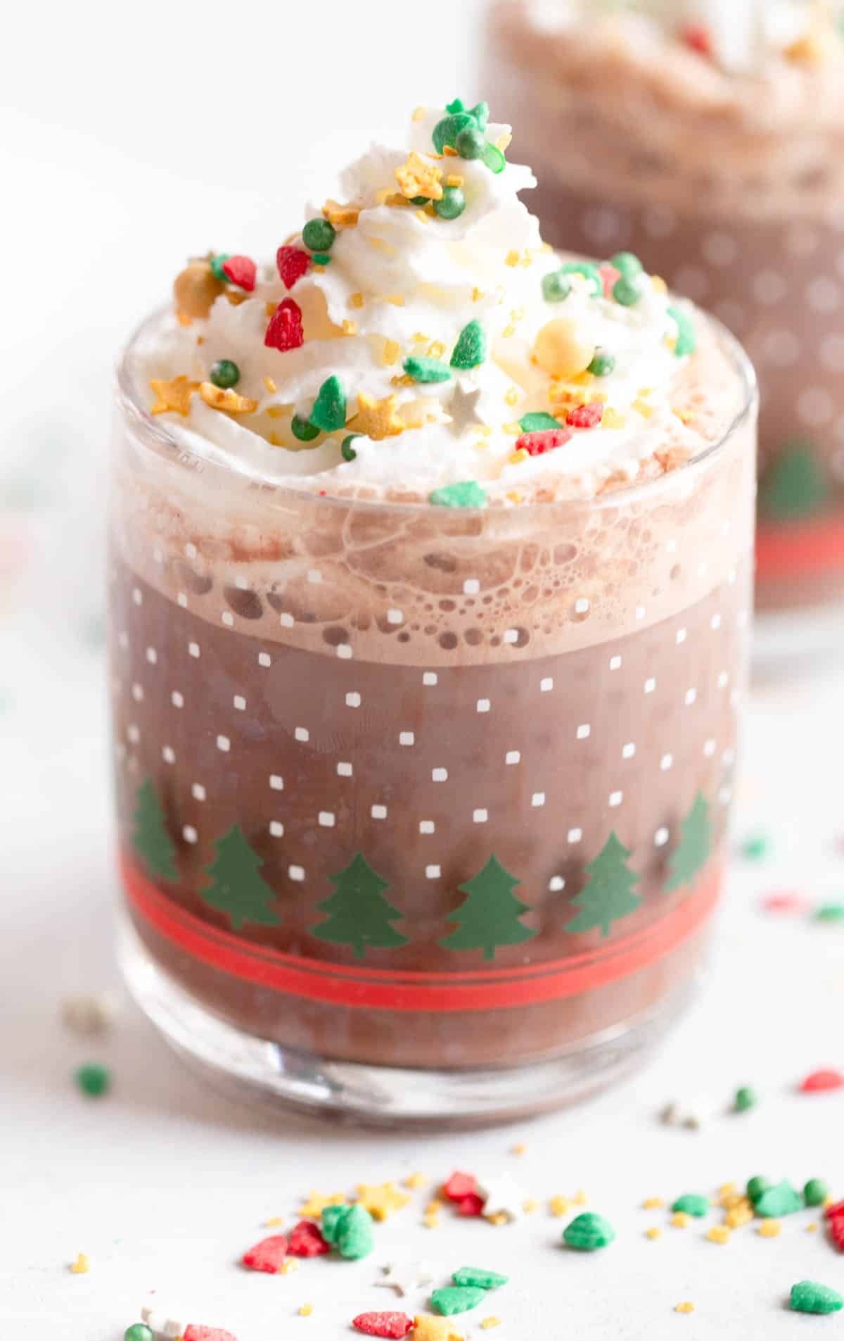 Homemade Hot Cocoa Mix, prepared, in a festive clear mug with Christmas trees on it. The hot cocoa has whipped cream on top with Christmas sprinkles on top and around the mug. 