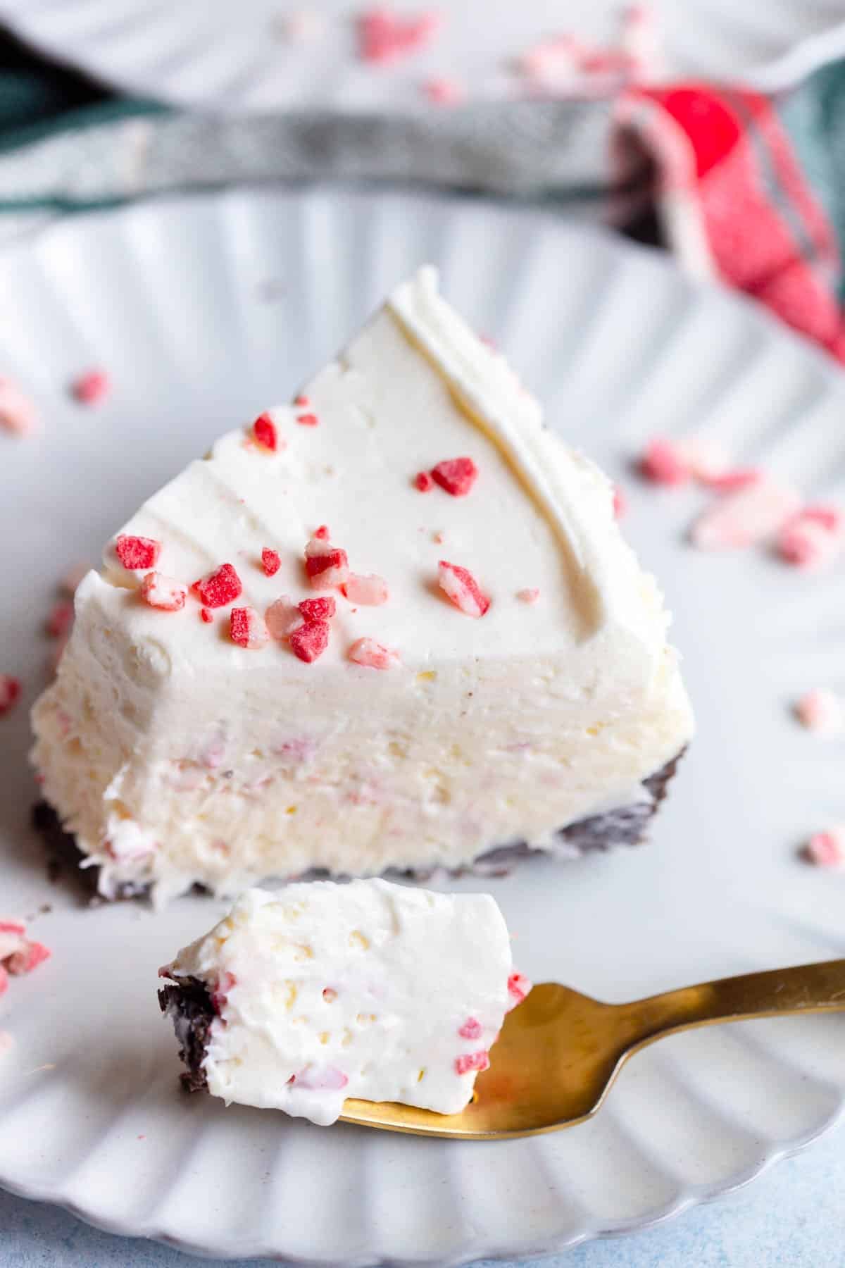 A slice of No Bake Peppermint Cheesecake on a scalloped plate with a gold fork that has taken a bite of cheesecake on it. There are Andes peppermint bark baking bits scattered around it and a gold fork in the background.