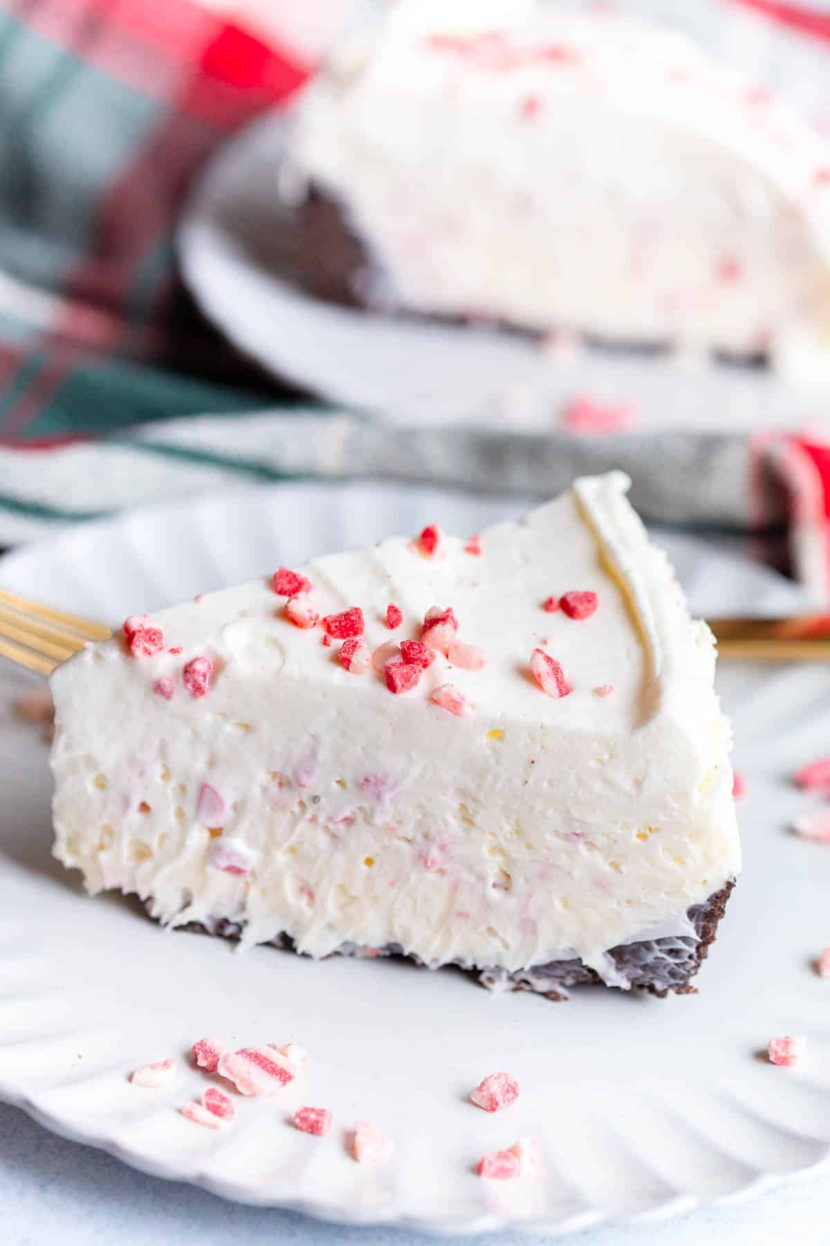 A slice of No Bake Peppermint Cheesecake on a scalloped plate with Andes peppermint bark baking bits scattered around it and a gold fork in the background.