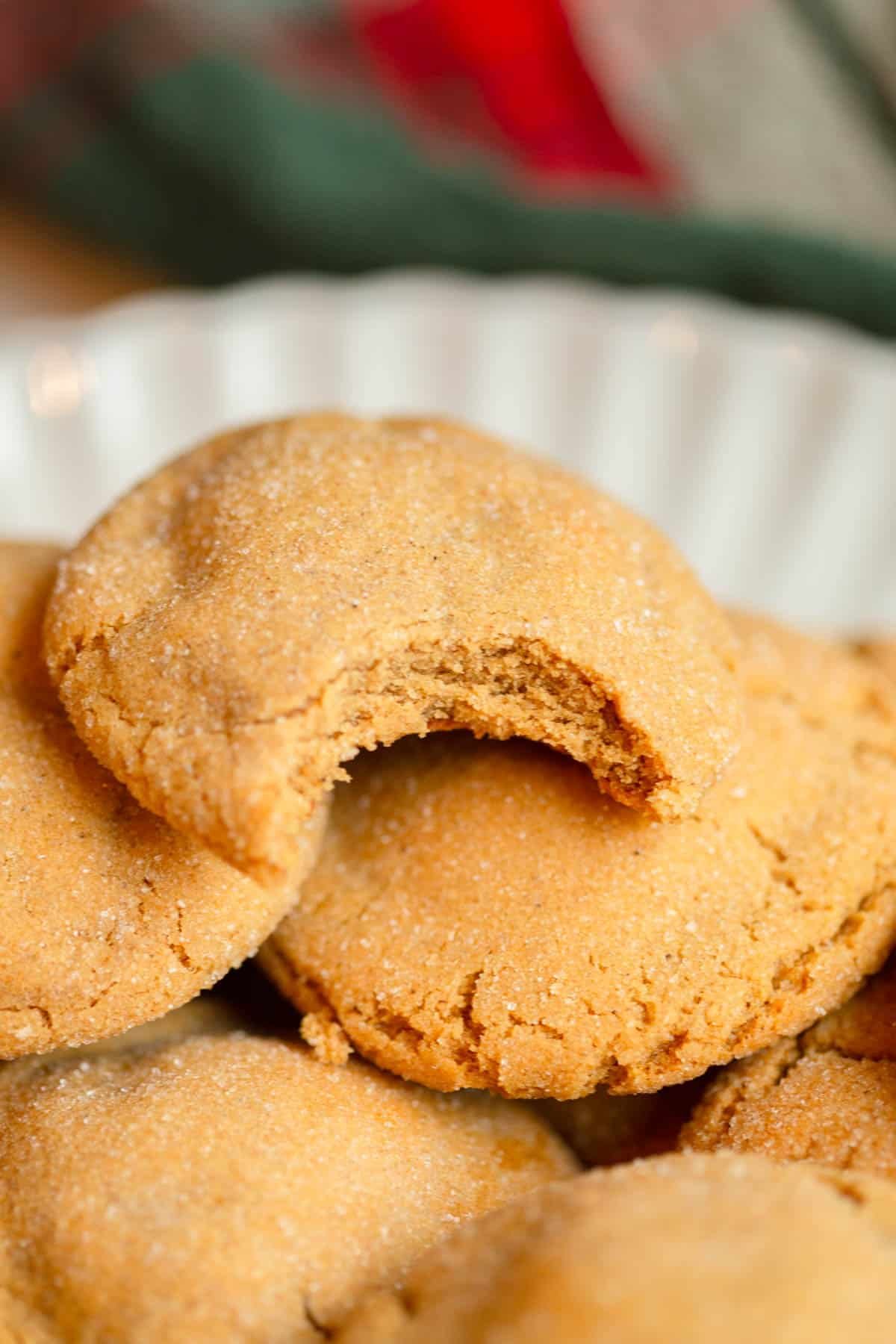 Chewy Ginger Cookies piled on a scalloped cream colored plate with a plaid red and green napkin in the background. One cookie has a bite taken out of it.