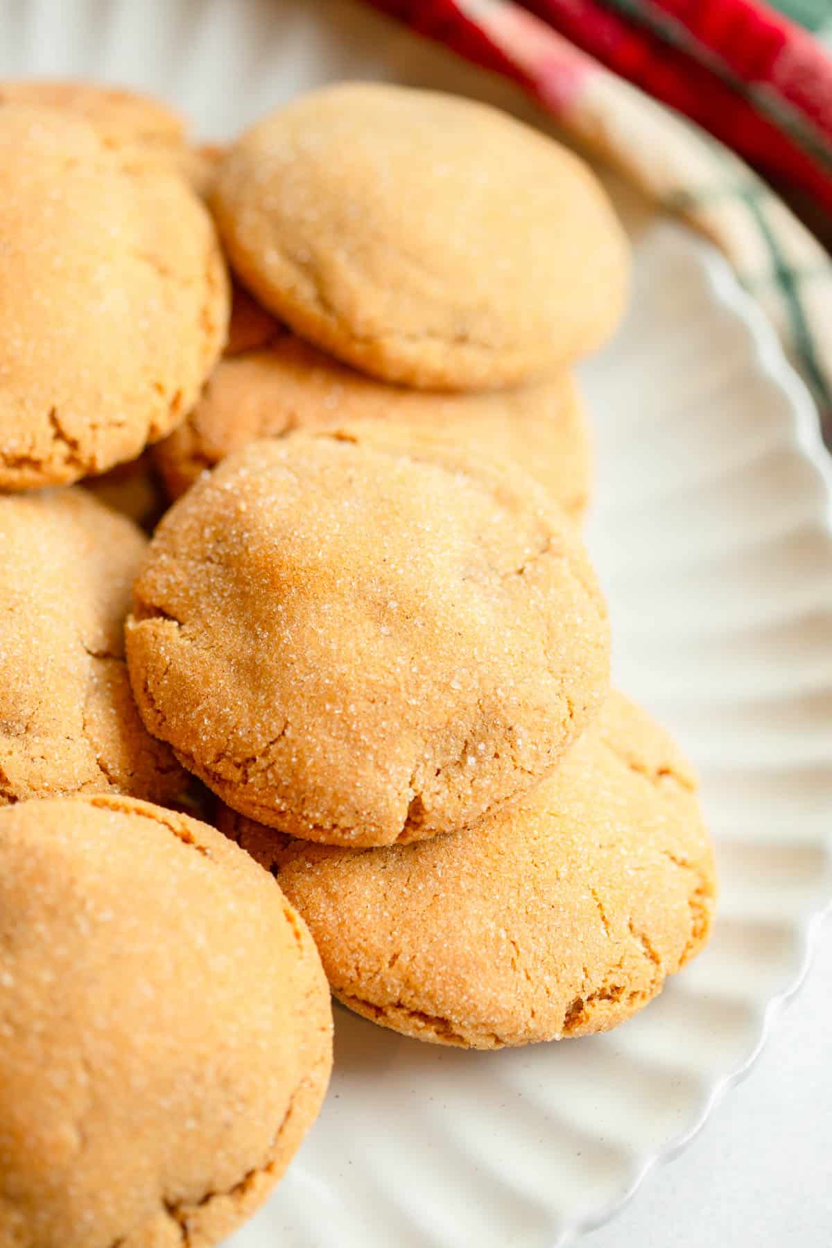 Chewy Ginger Cookies piled on a scalloped cream colored plate with a plaid red and green napkin in the background. 