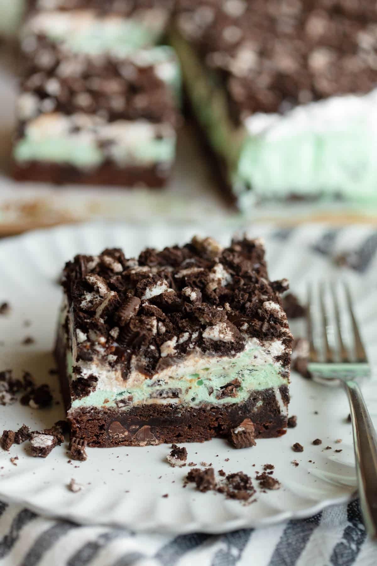 A Mint Brownie Ice Cream Bar on an off white scalloped plate with a fork next to it. There are chopped Oreos scattered around the bar on the plate. 