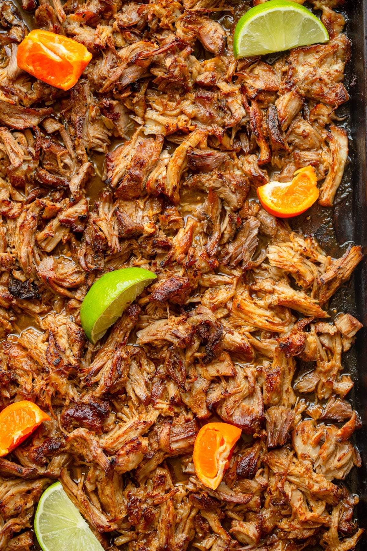 Pork carnitas meat on a baking sheet with wedges of lime and oranges.