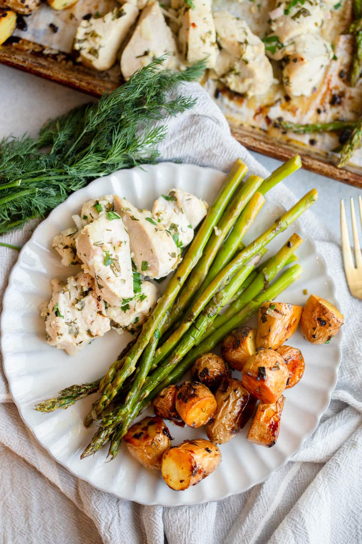 Sheet Pan Lemon Chicken and Potatoes on a scalloped cream colored plate with fresh dill and the baking sheet next to the plate. 