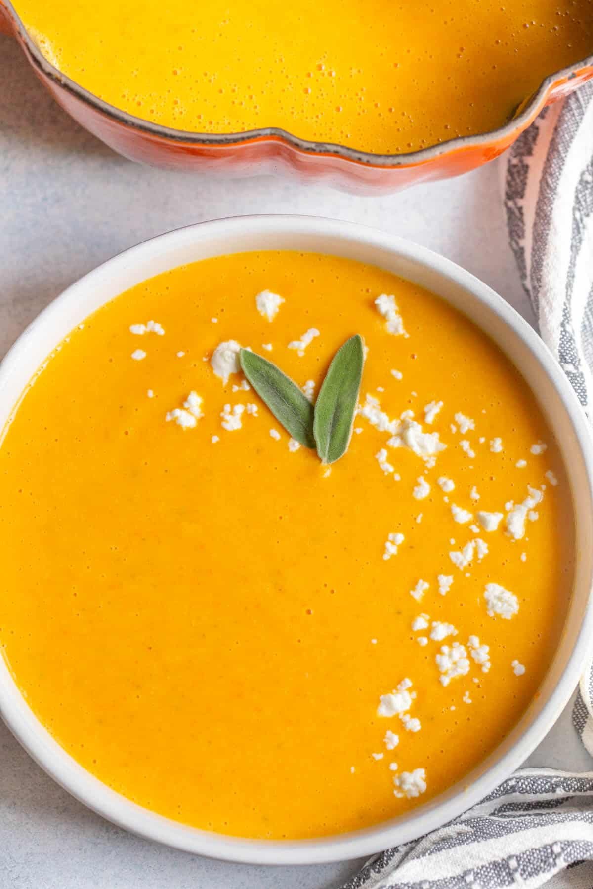 Boursin Butternut Squash Soup in a white bowl with crumbled goat cheese sprinkled on top and two sage leaves as garnish. 