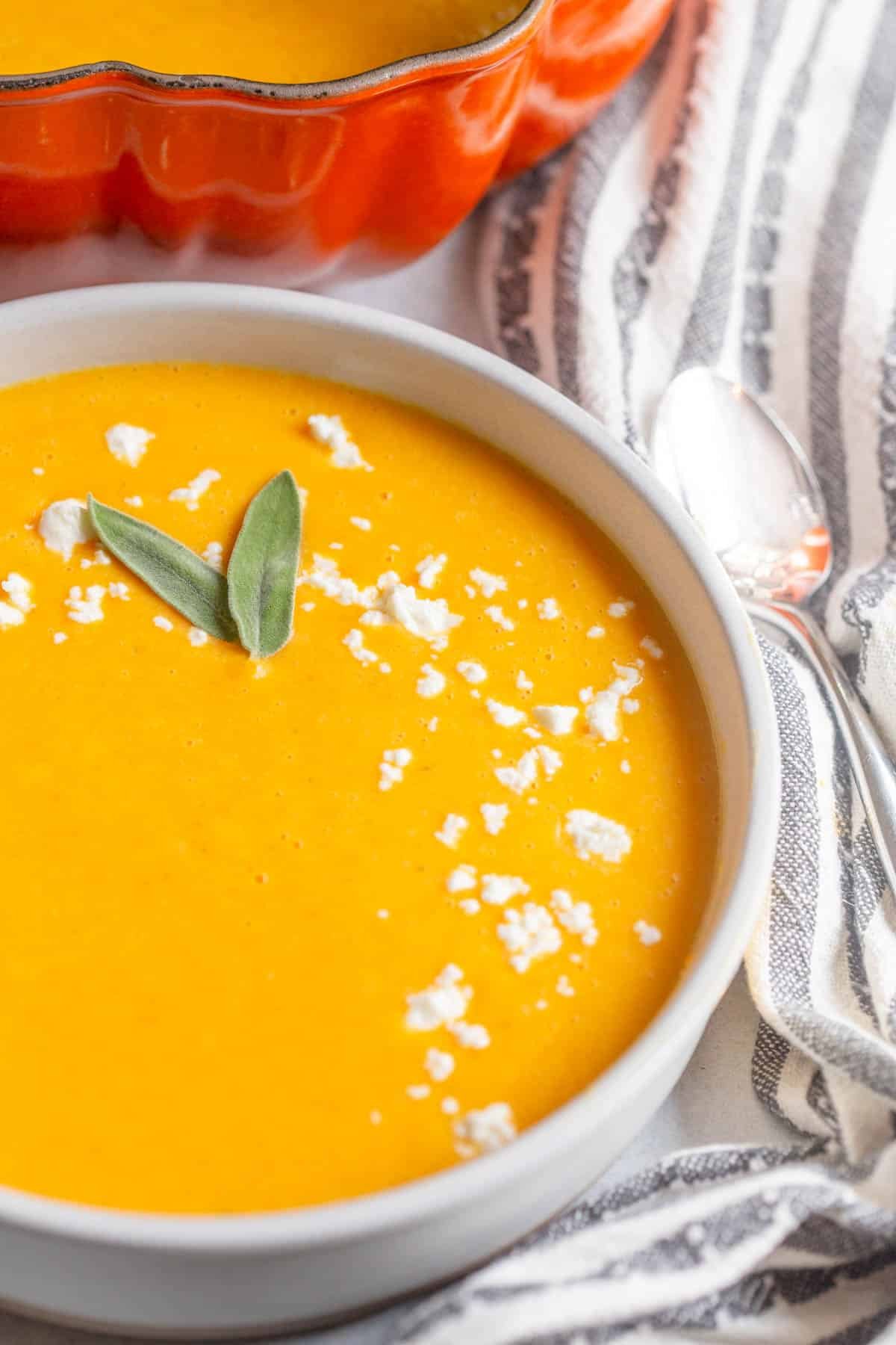 Boursin Butternut Squash Soup in a white bowl with a spoon next to it, with crumbled goat cheese sprinkled on top and two sage leaves as garnish. 