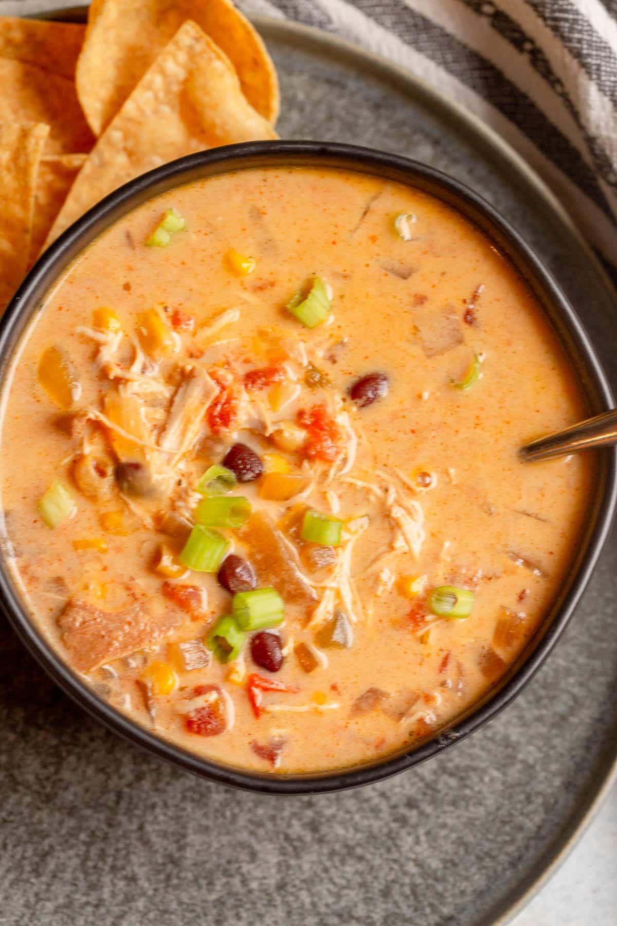 Creamy Chicken Tortilla Soup in a black bowl which is sitting on a dark gray plate. There is a black and white striped napkin next to the plate and a silver spoon resting in the bowl of soup.
