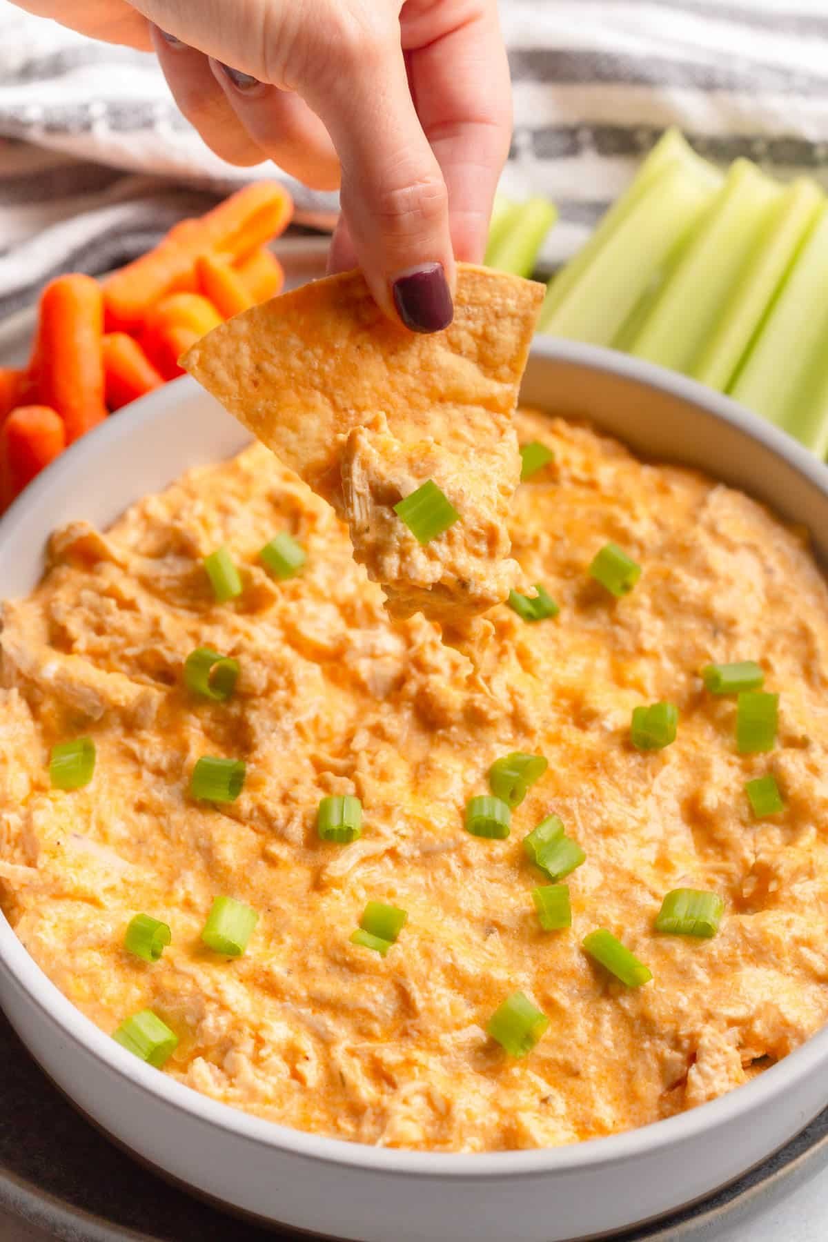 High Protein Buffalo Chicken Dip in a shallow cream colored bowl topped with diced green onions, with celery sticks and baby carrots in the background, and a hand with a tortilla chip topped with the dip.