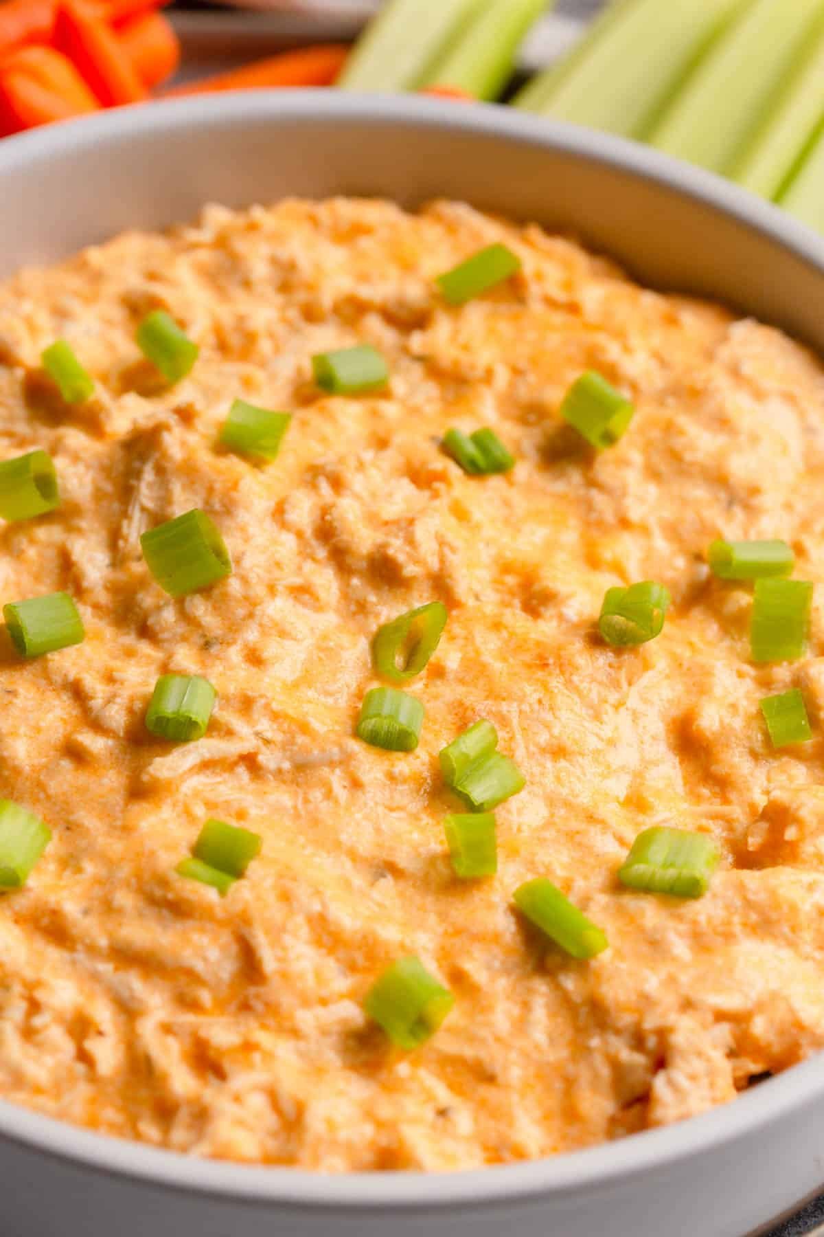High Protein Buffalo Chicken Dip in a shallow cream colored bowl topped with diced green onions, with celery sticks and baby carrots in the background. 