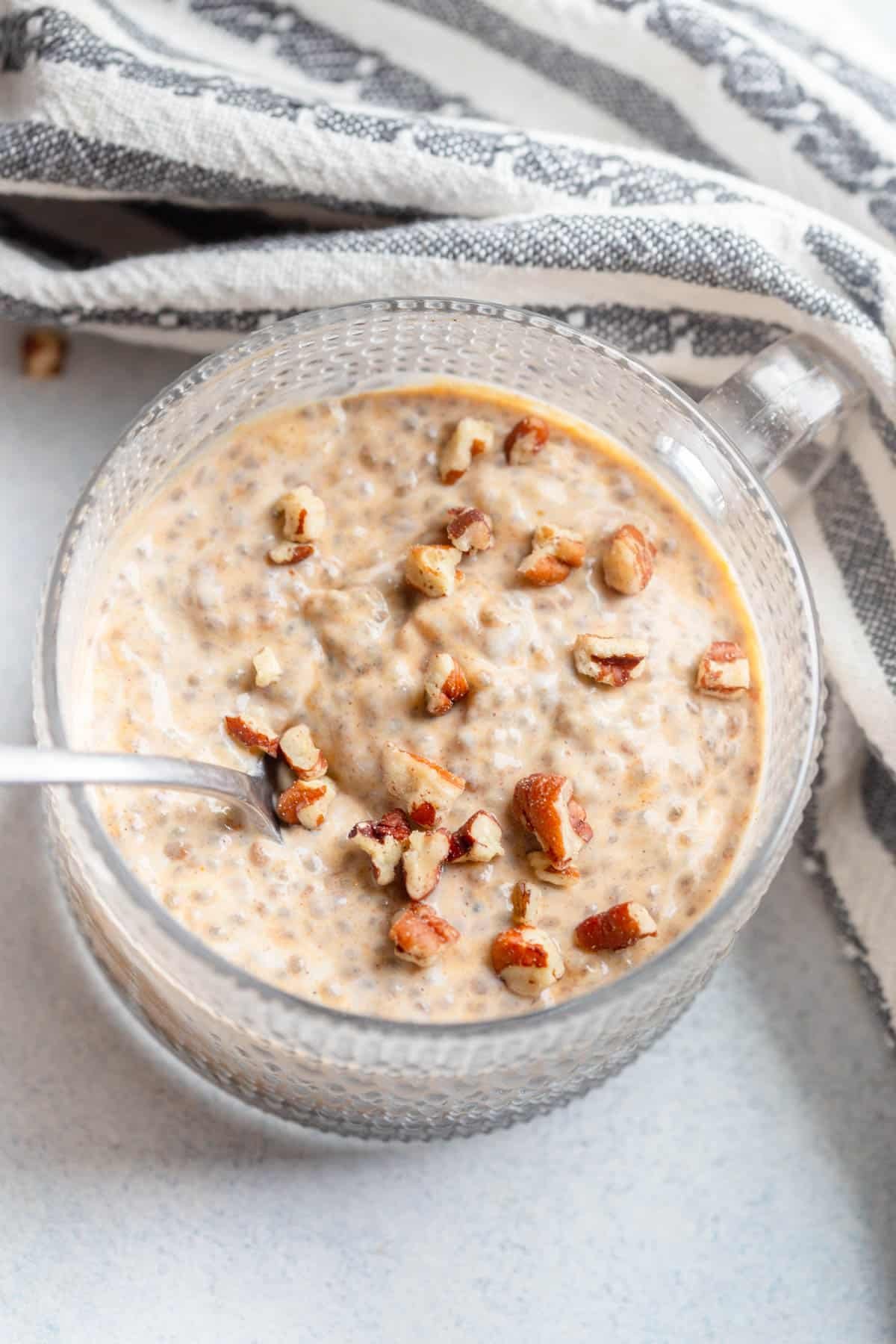 Pumpkin Chia Seeding Pudding in a glass mug with a spoon in it. There are chopped pecans topping the chia seed pudding and a black and white striped towel next to it. 